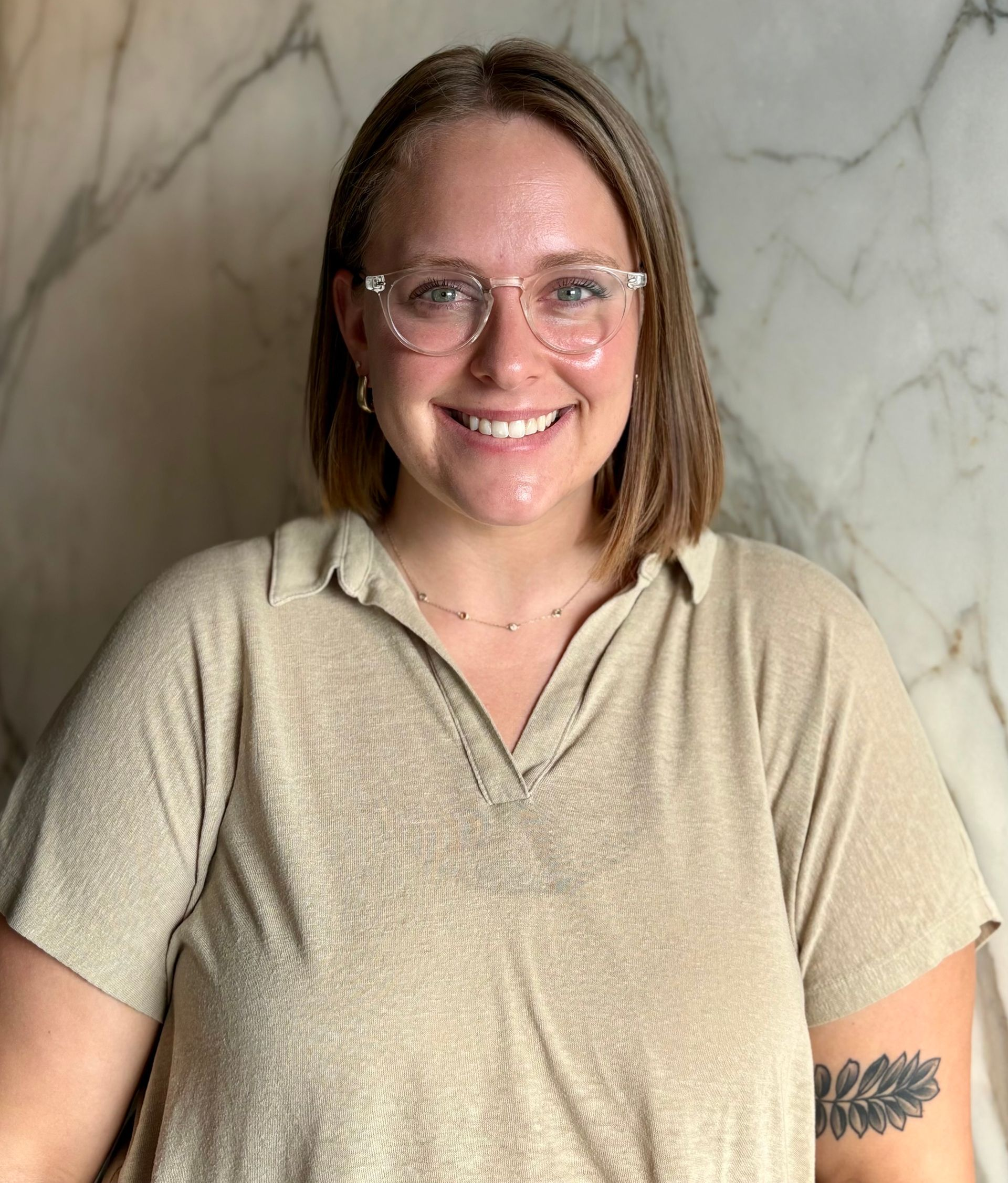 Woman with glasses smiles; wearing a tan shirt, stands in front of a marble wall, and has a tattoo on her arm.