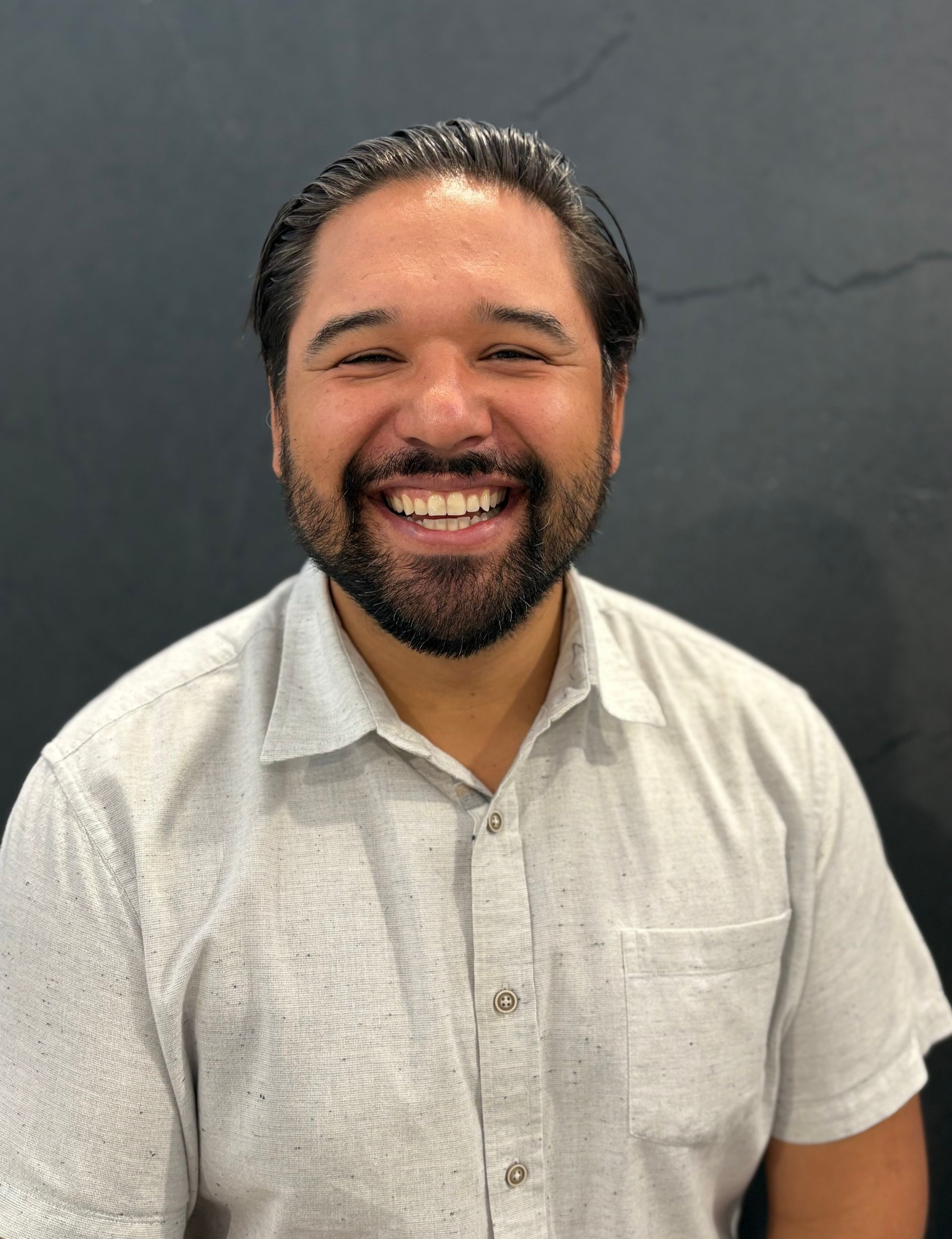 Man smiling, wearing a light-colored button-up shirt, standing in front of a dark background.