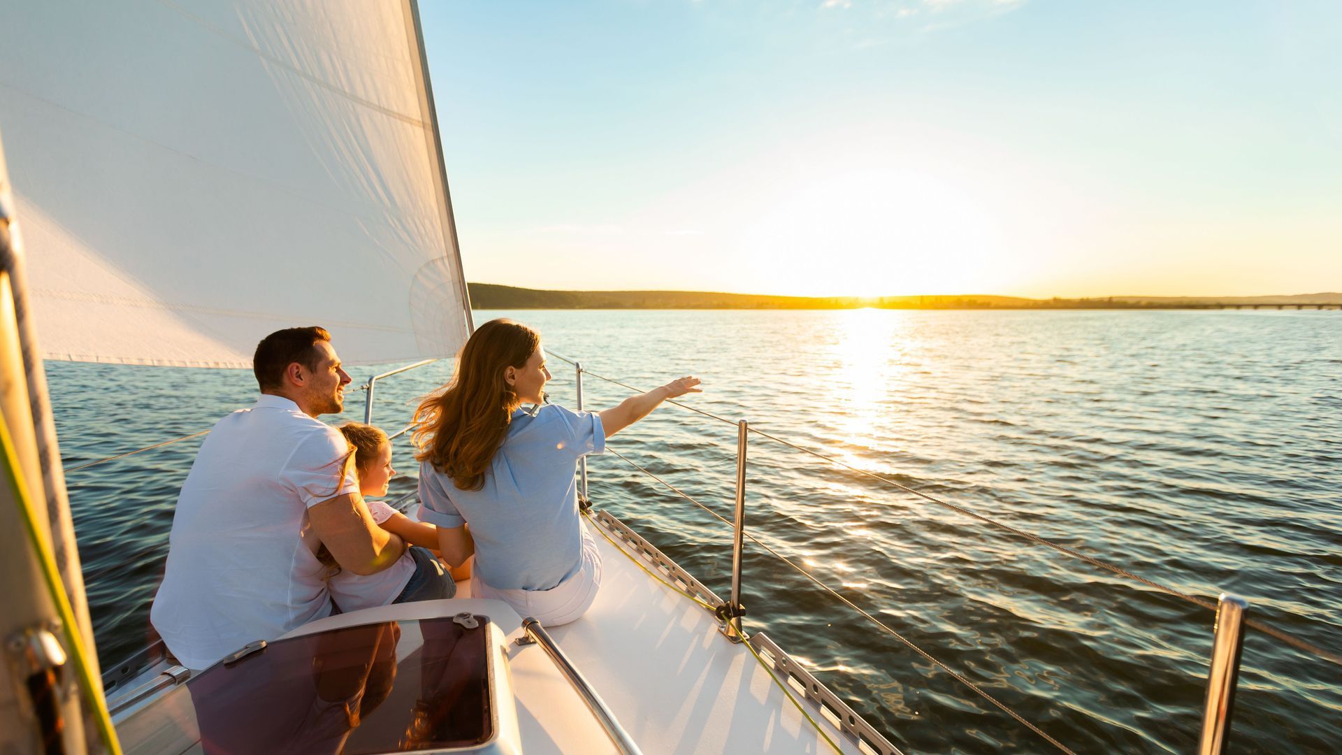 A man and a woman are sitting on a sailboat looking at the sunset.