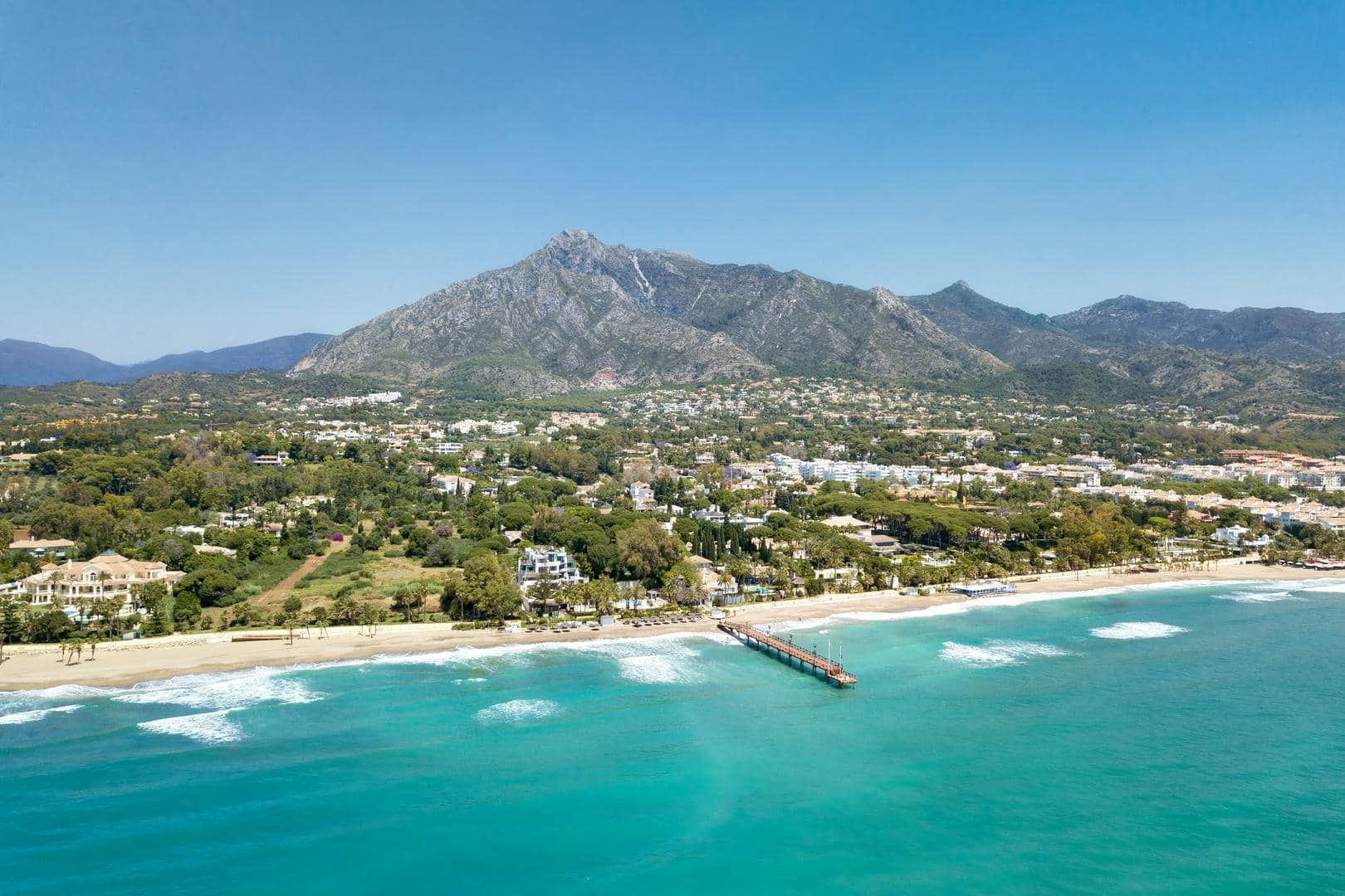 An aerial view of a beach with mountains in the background and a pier in the foreground.