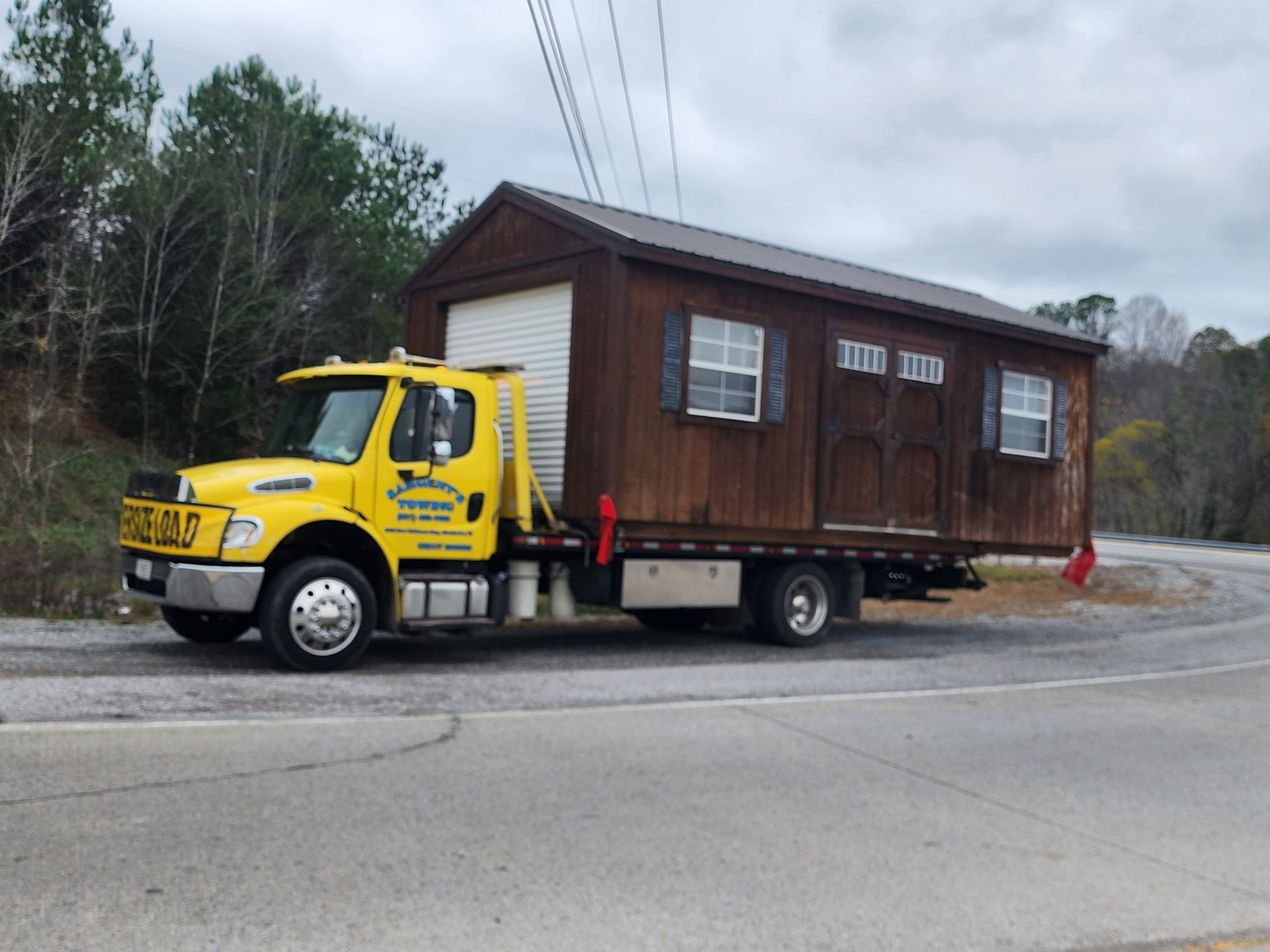 Yellow truck transporting a brown shed on a roadside.