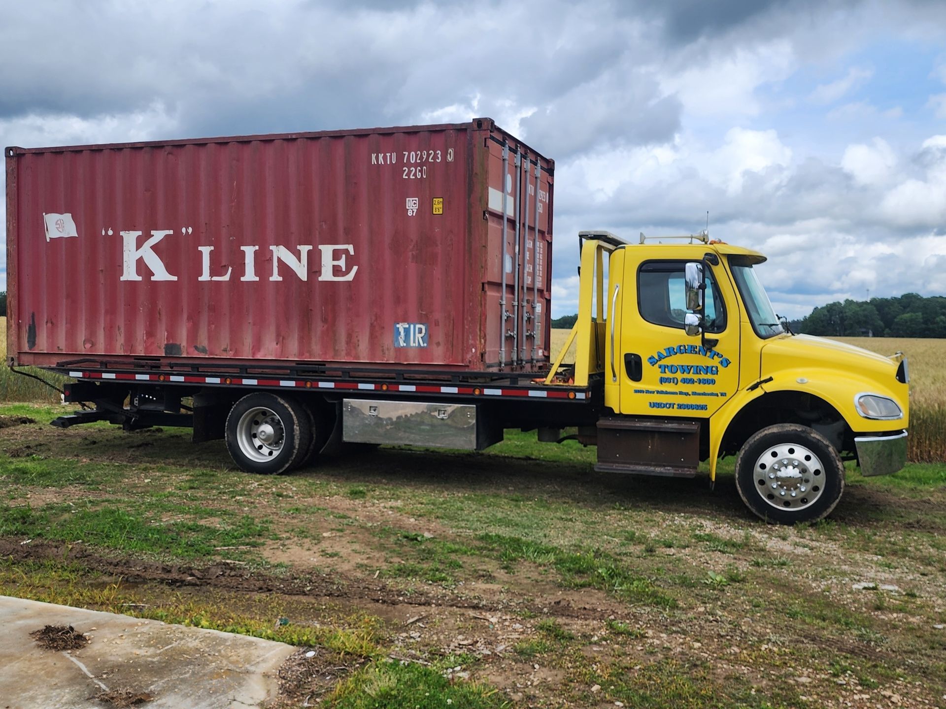 Yellow flatbed truck carrying a red K-Line shipping container on a grassy field.