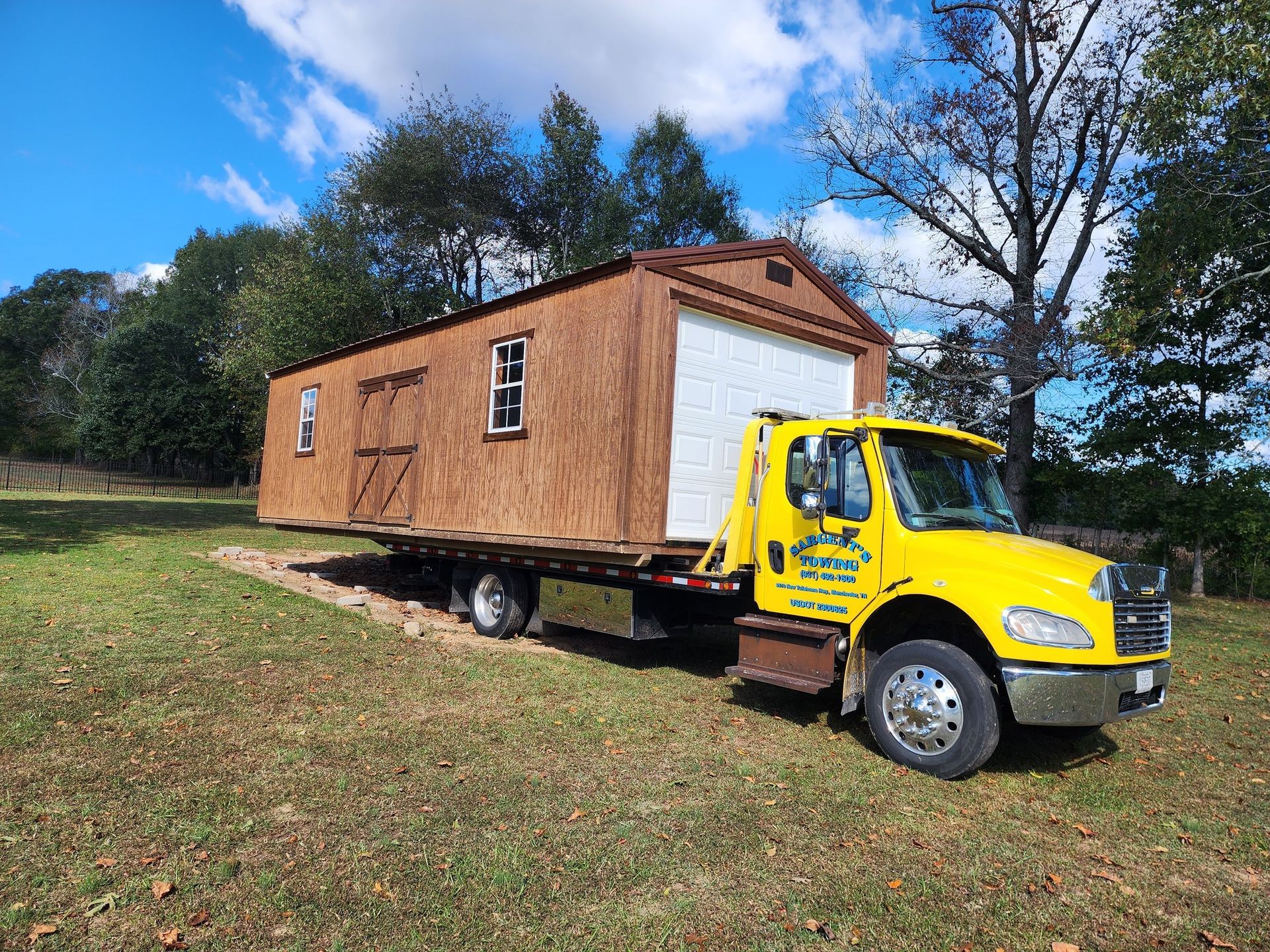Yellow truck transporting a brown shed on a flatbed trailer, set on a grassy field under a blue sky.