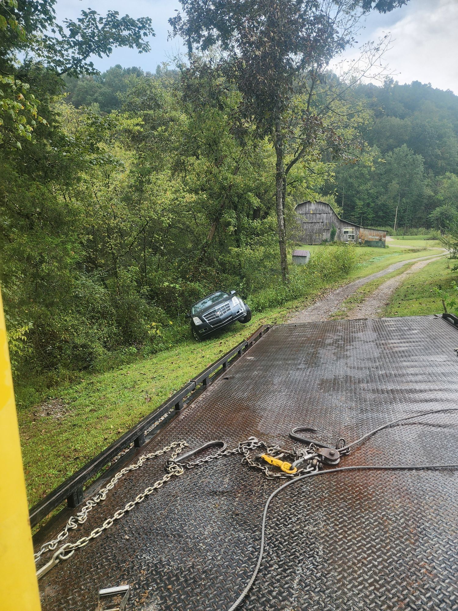 A car being towed from a grassy area next to a gravel road, with trees and a building in the background.