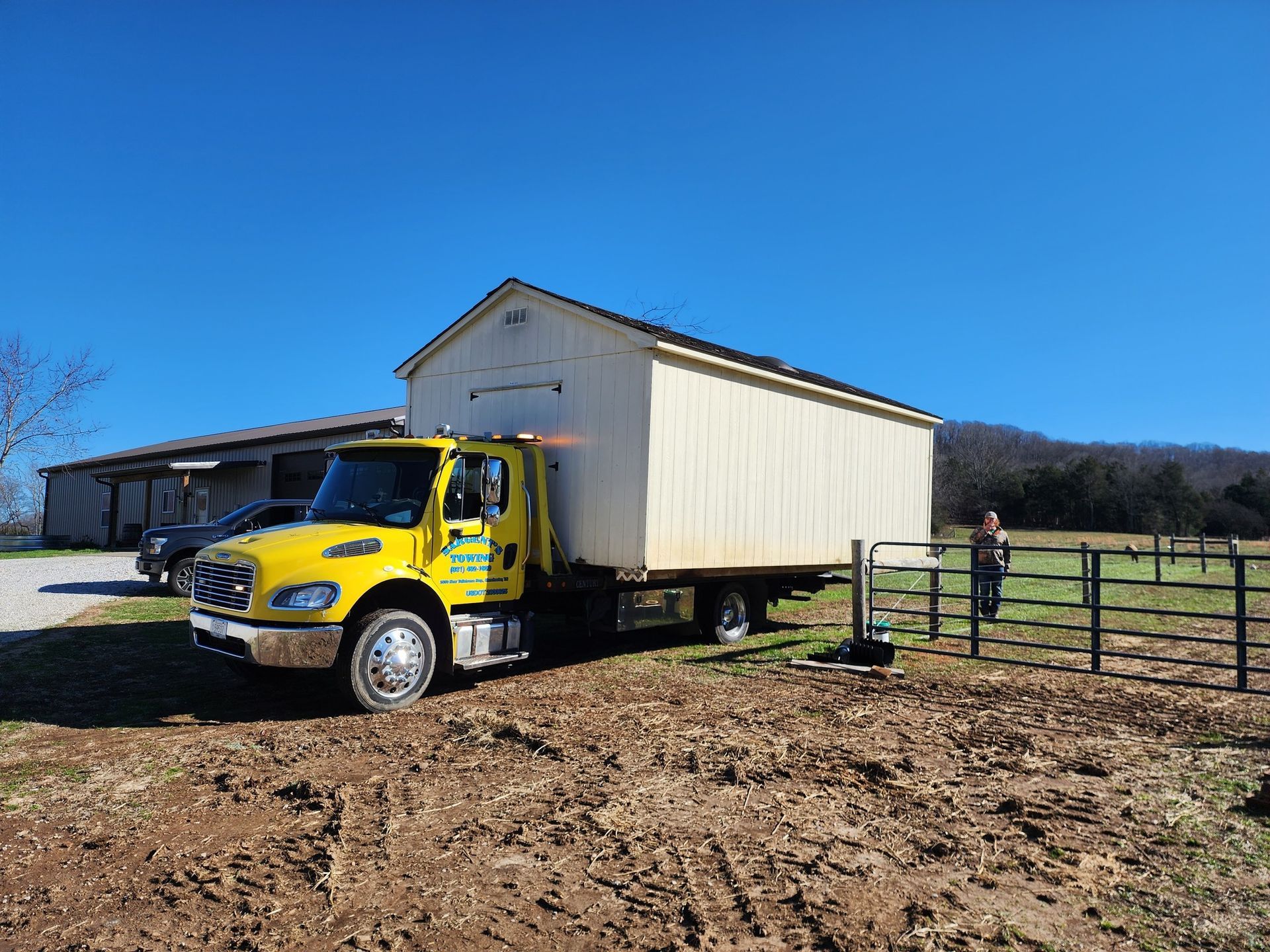 Yellow truck transporting a white shed in a field, under a blue sky. A person stands nearby.