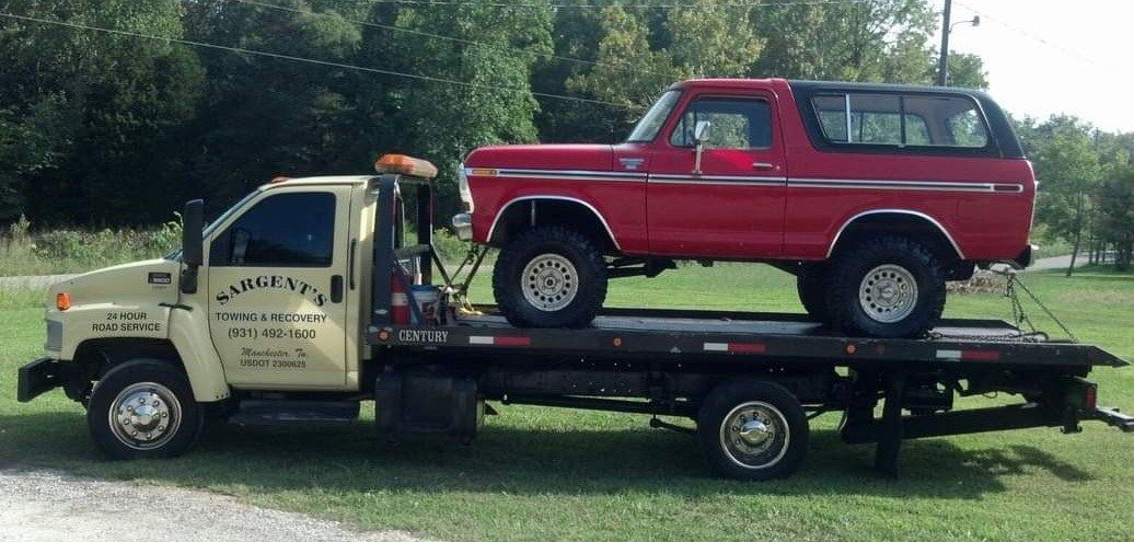 A red Bronco SUV being towed on a flatbed tow truck. The truck is beige. Green trees in background.