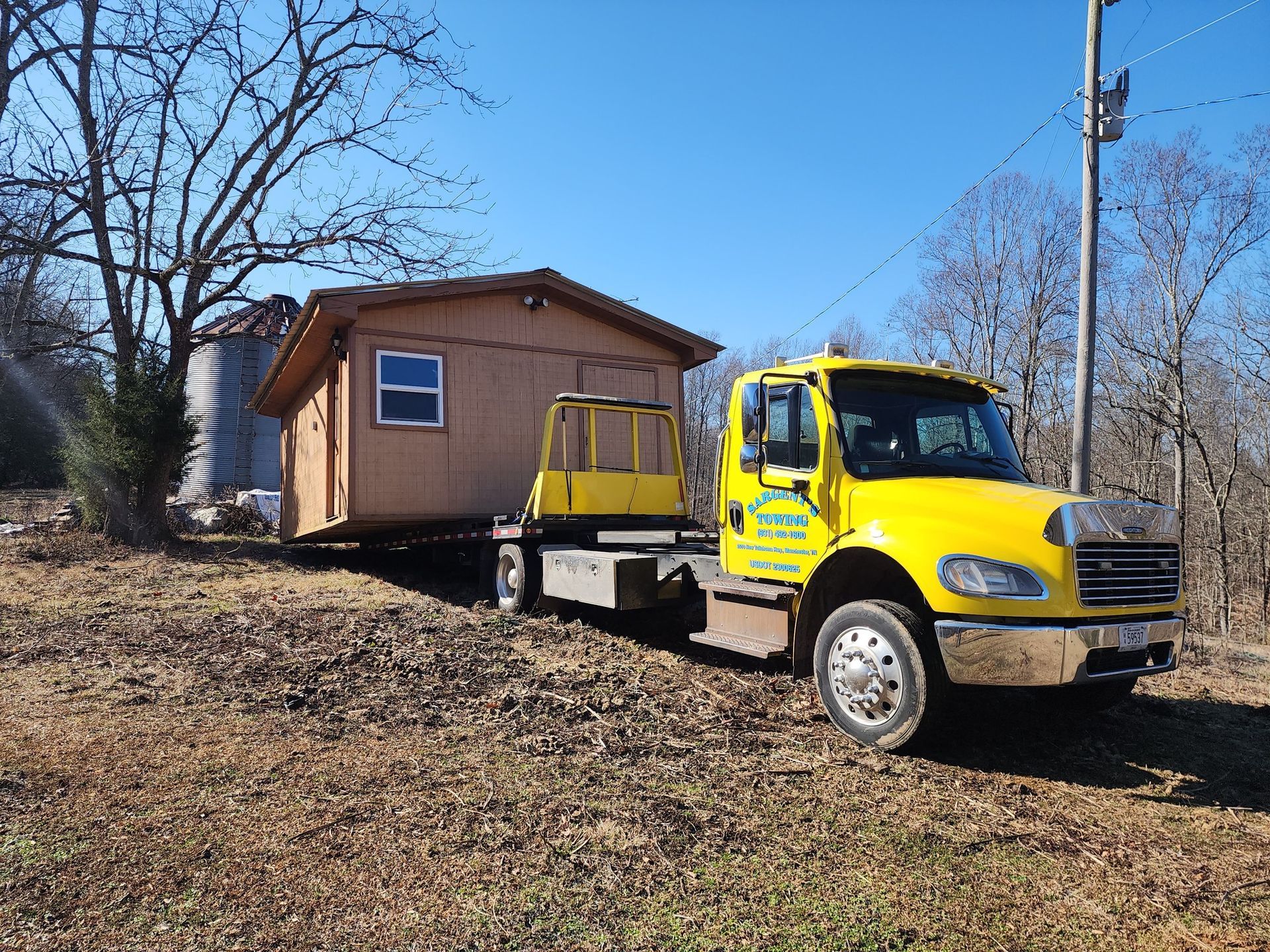 Yellow truck hauling a small wooden shed on a sunny day.