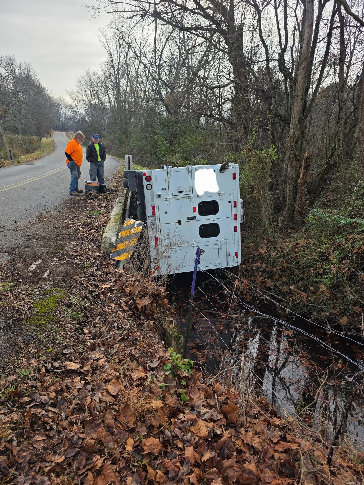 Two men beside a utility truck, angled into a ditch beside a road. Trees surround.