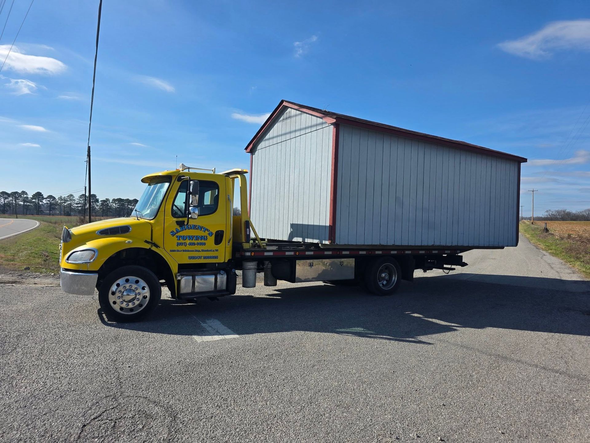 Yellow tow truck transporting a gray shed on a paved road under a blue sky.