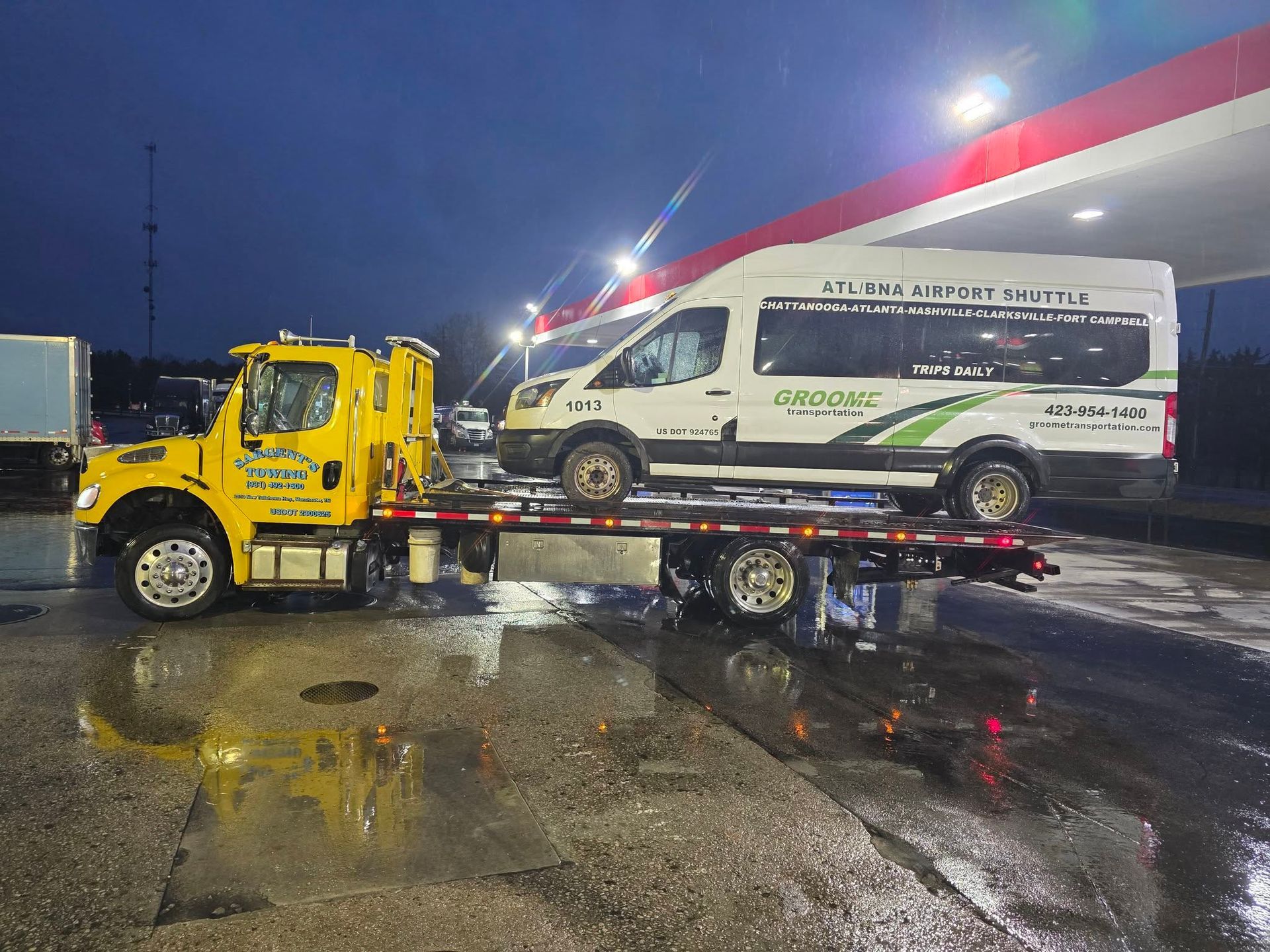 Yellow tow truck towing a white passenger van at a gas station at night. Wet pavement.