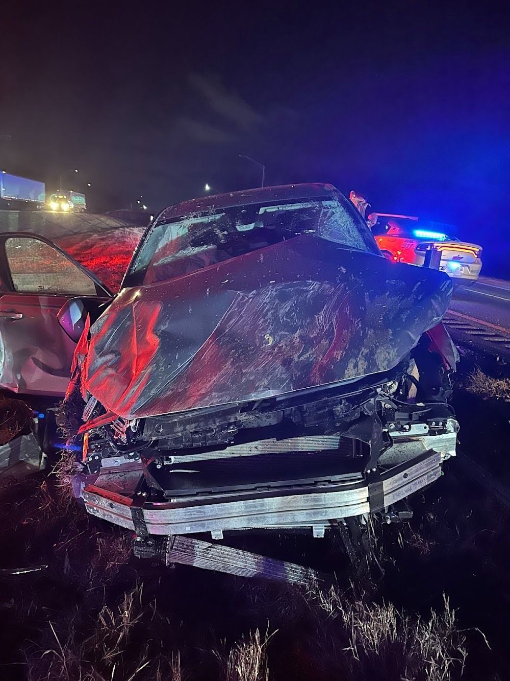 Severely damaged car at night after a collision, emergency lights visible in the background.