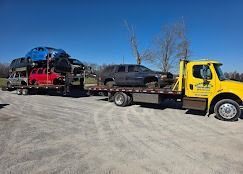 Yellow tow truck hauling a trailer loaded with several wrecked cars on a sunny day.