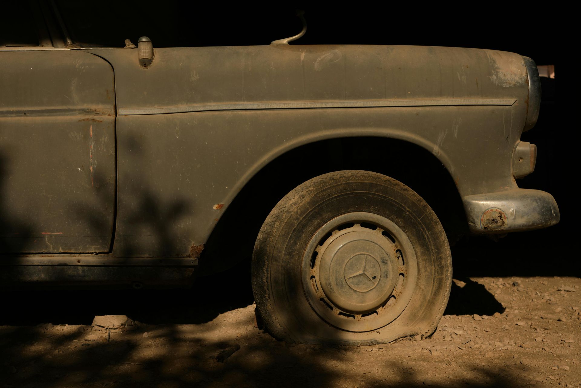 Old, weathered gray car with flat tire on dirt ground; shadows cast.
