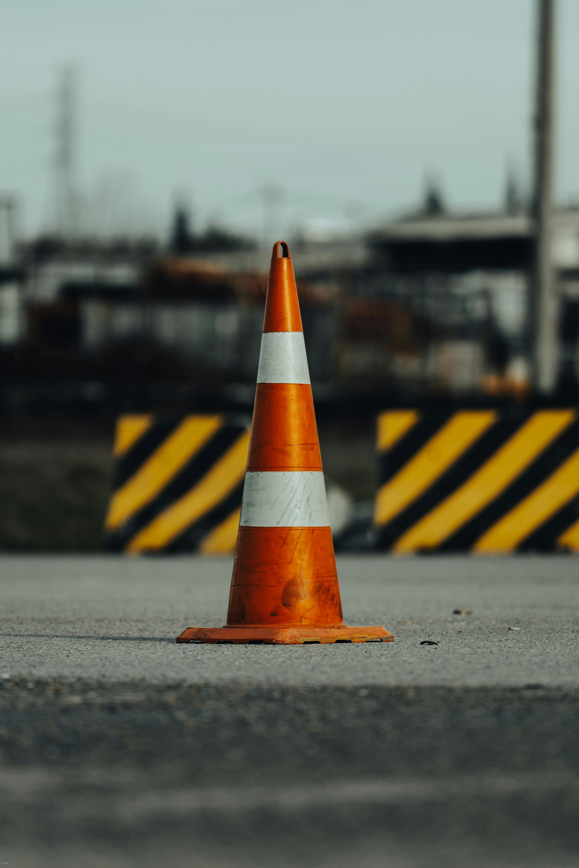 Orange and white traffic cone on asphalt, with yellow and black barricades in the background.