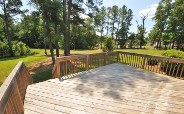 A photo of a large deck in a house's backyard. The house's backyard is wide open and has a lot of trees in the distance. The deck looks beautiful but it is empty with no furniture or decor on it. 