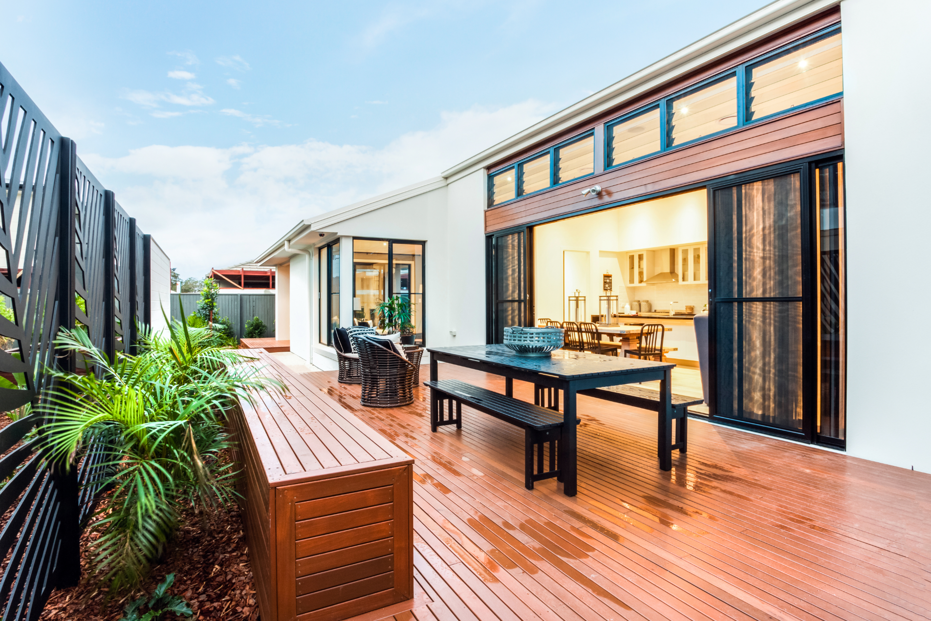 A beautiful photo of a wooden deck/patio that is built on the back of a modern house. The deck is filled with furniture and plants.