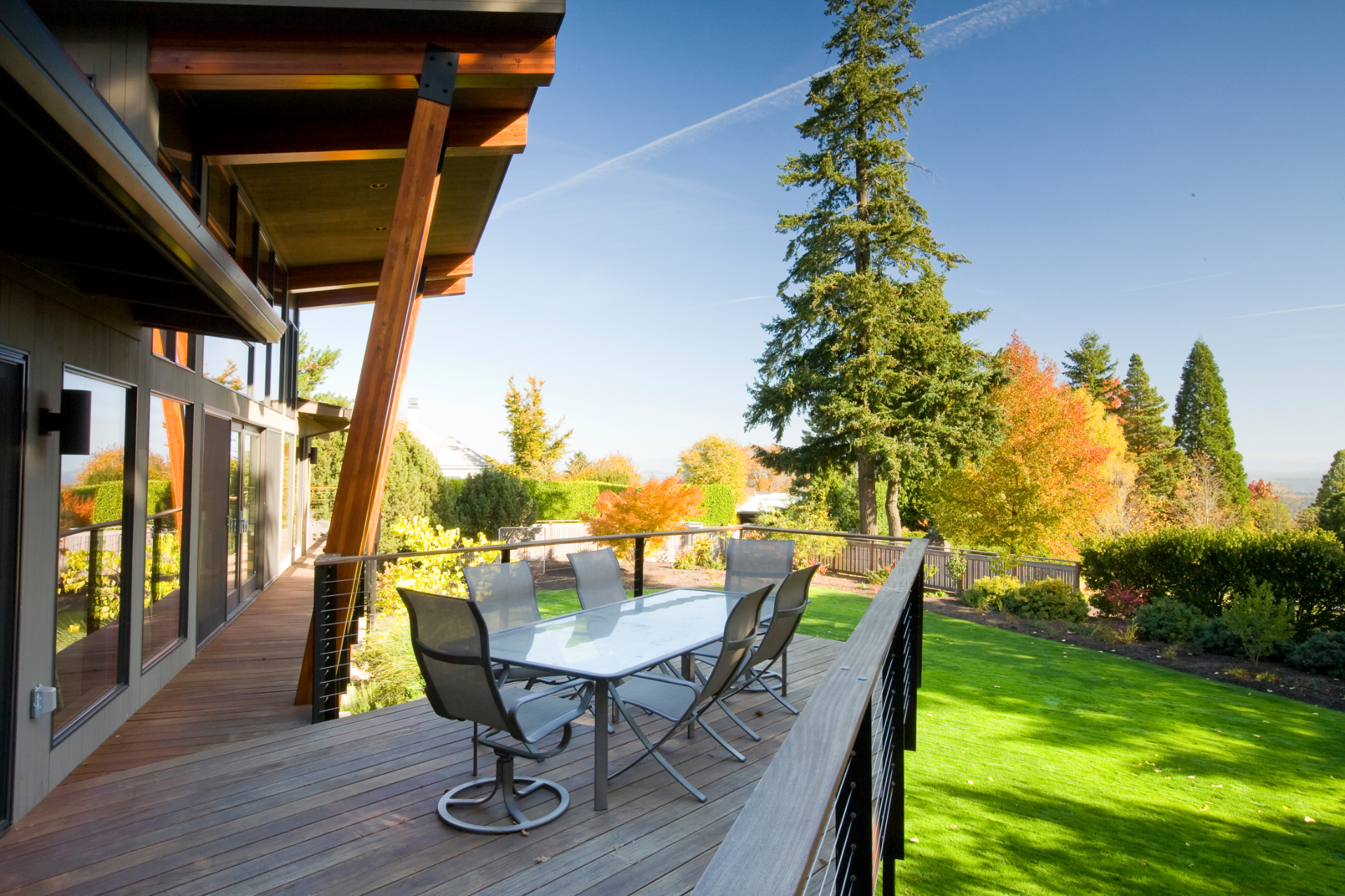 A beautiful photo of a second-level deck that is built on the back of a very nice house. The deck overlooks a beautiful yard with green trees and grass. The deck has a table with chairs on it.