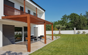 A photo of a pergola installed onto the back of a house. The pergola offers some shade to the furniture nestled under it. It is a sunny day with no clouds in the sky. The pergola sits on top of a patio and extends right up to the edge of the patio, where it meets the grass.