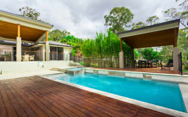 A stunning photo of a deck surrounding a pool and hot tub in the backyard of a modern home. The pool deck also encompasses a pergola just beyond the pool to offer shade and protection from the sun. 
