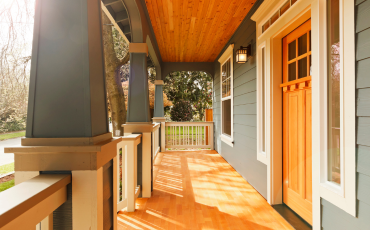 A photo of a small but cozy porch in front of a nice house. The sun is shining brightly on the porch. The porch is a mix of white railings, blue pillars, and tan floors and ceilings. 