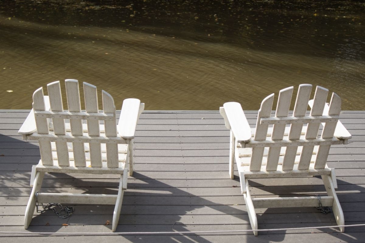 Two white Adirondack chairs sit facing a calm body of water on a composite grey deck in Stockton, CA. The chairs' slightly weathered texture adds character, suggesting many peaceful hours spent enjoying the view. Fallen leaves dot the deck, indicating the autumn season. The water's surface is gently rippled, reflecting a serene environment. This setup invites relaxation and contemplation, a quiet corner perfect for unwinding in the presence of nature.