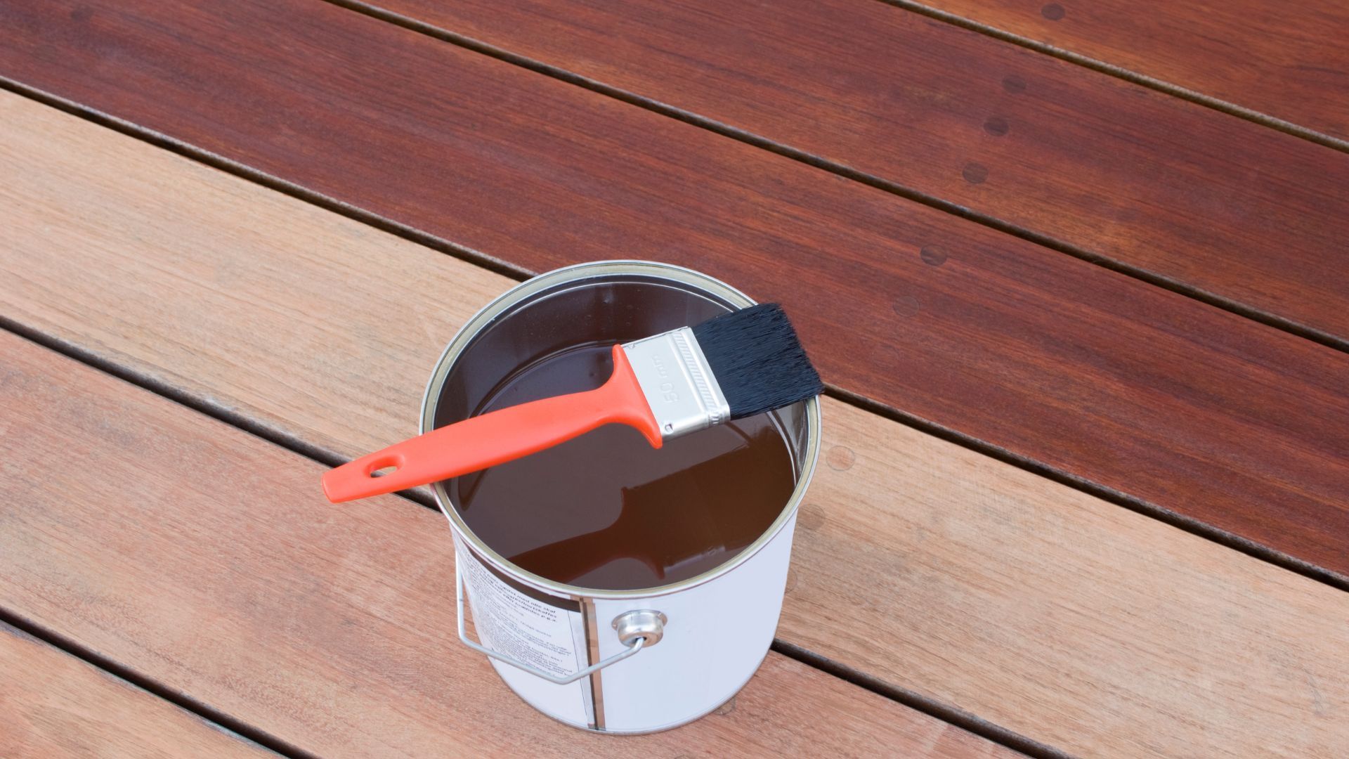 Can of wood stain with a brush on a partially stained deck in Stockton, CA, showing color contrast.