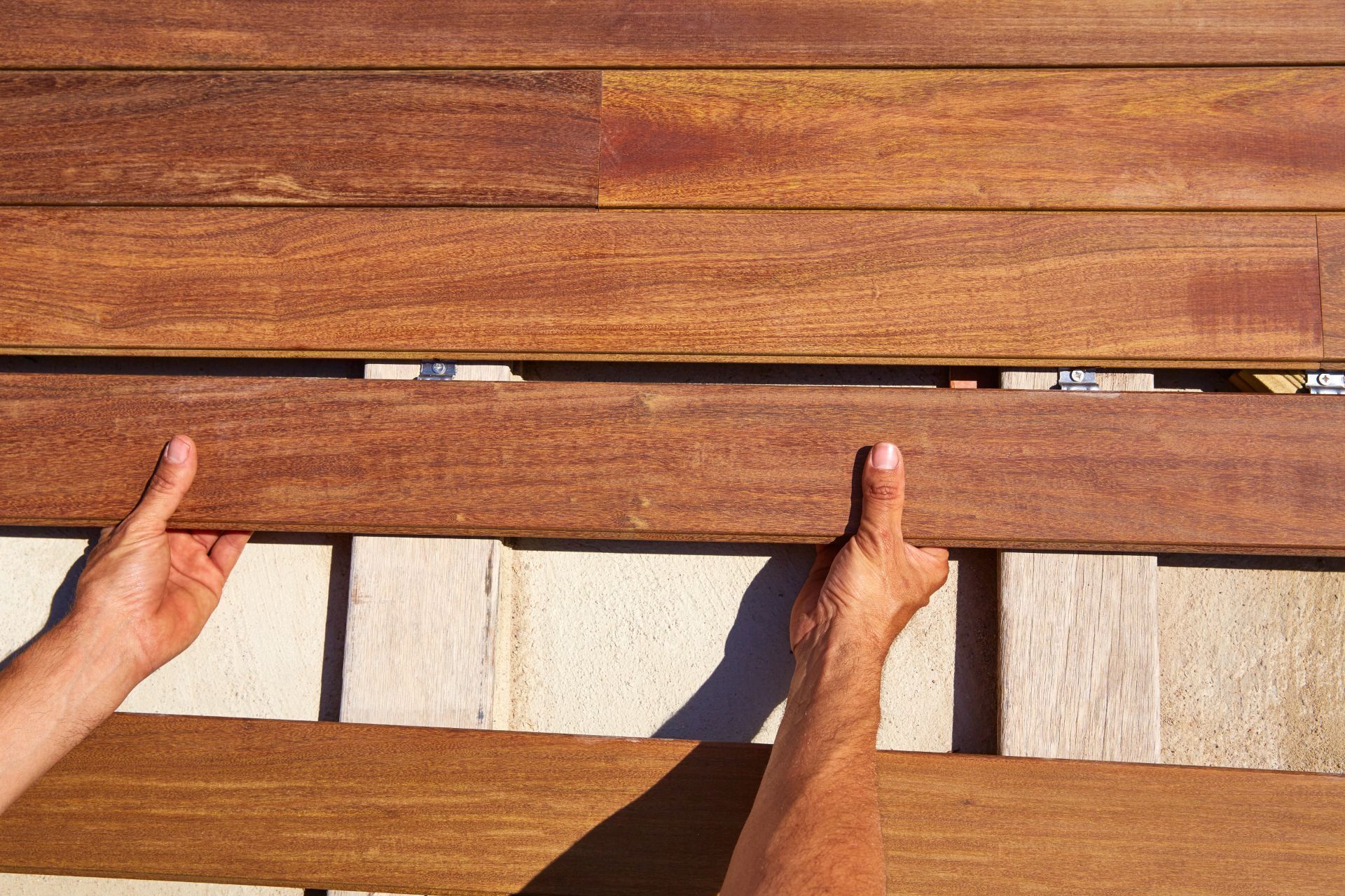 A person's hands are shown installing wooden deck boards on a sunny day in Stockton, California. The top board is being aligned with the others, which have a warm, reddish-brown stain highlighting the wood grain. Visible brackets and screws secure the boards in place, reflecting an in-progress deck construction with a focus on craftsmanship and detail.