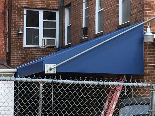 Blue awning attached to a brick building, supported by a cable and post above a chain-link fence.