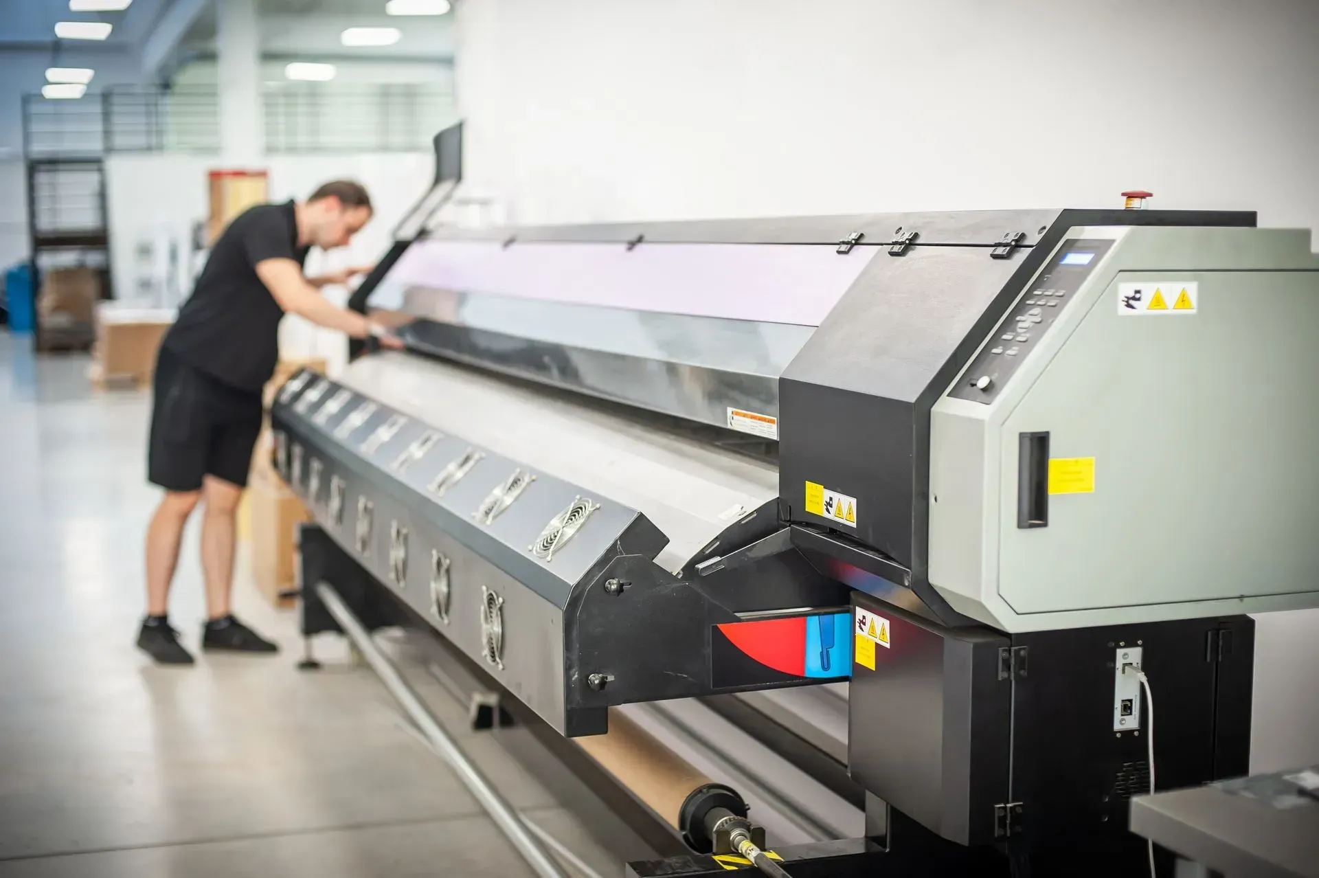 Man in black shorts operates a large-format printer in a bright warehouse setting.