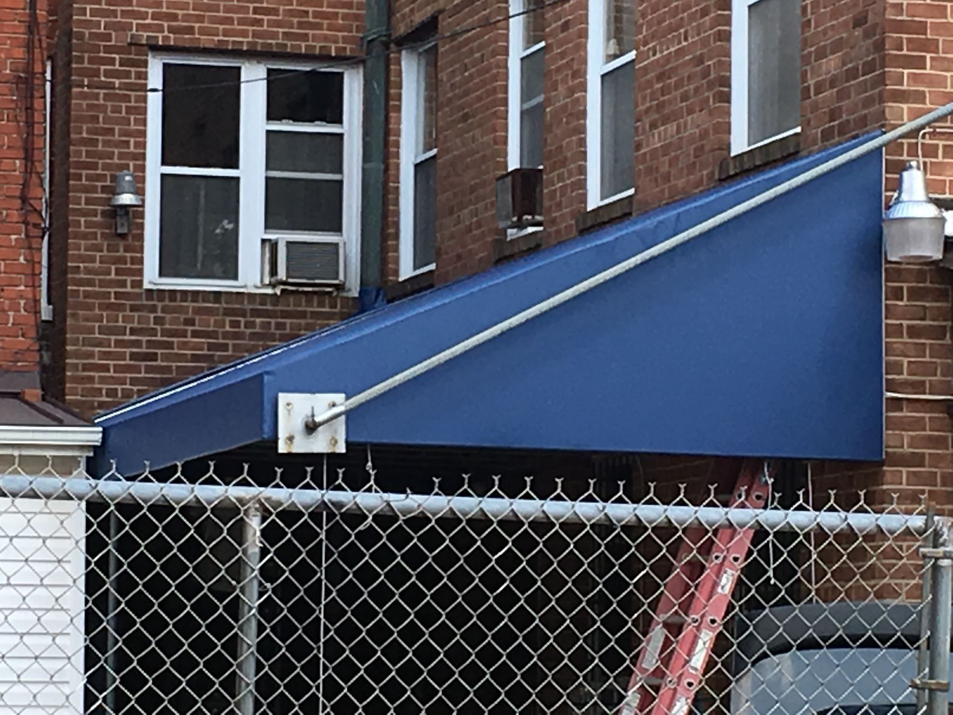 Blue awning angled against brick building. Chain link fence in front.