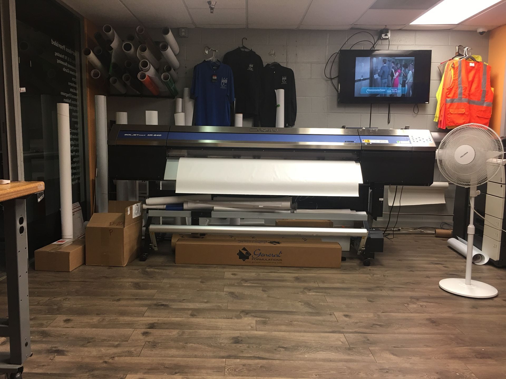 A large format printer in a workshop setting, with rolls of paper, clothes, and a TV.