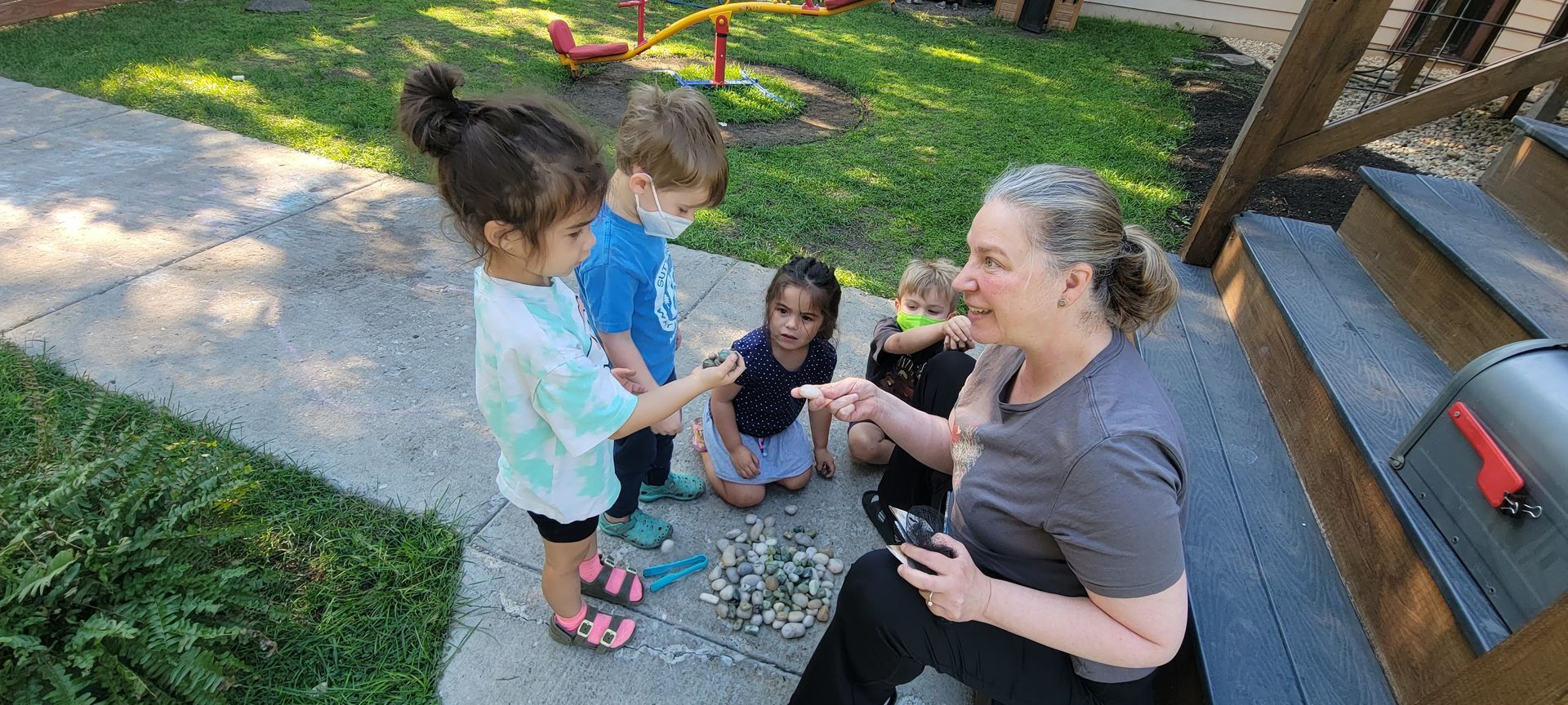 An adult woman sits on the steps of a backyard porch. A group of young children are sitting and standing near her. They are all examining a pile of small rocks and stones on the ground. Play equipment is in the background.