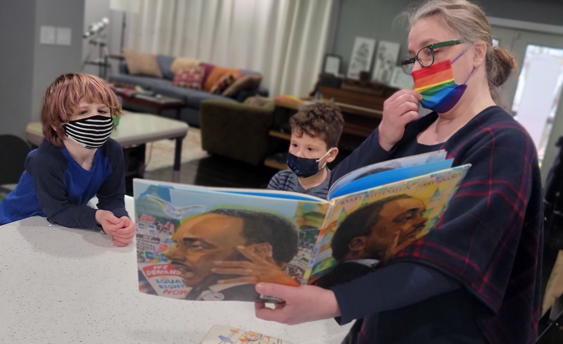 A teacher reads a storyboo, to two small children. All are wearing facemasks. The teacher is showing a picutre from the open book to the children.