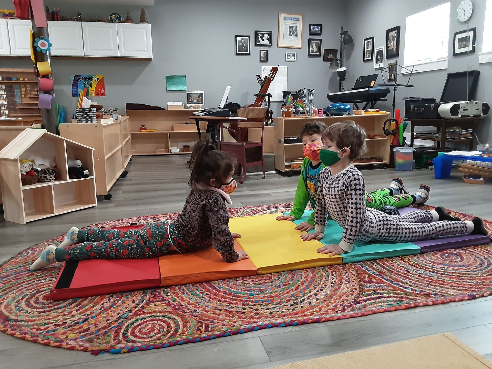 Three children are on the floor of a preschool setting, and are all doing the cobra yoga pose on a rainbow colored floormat.