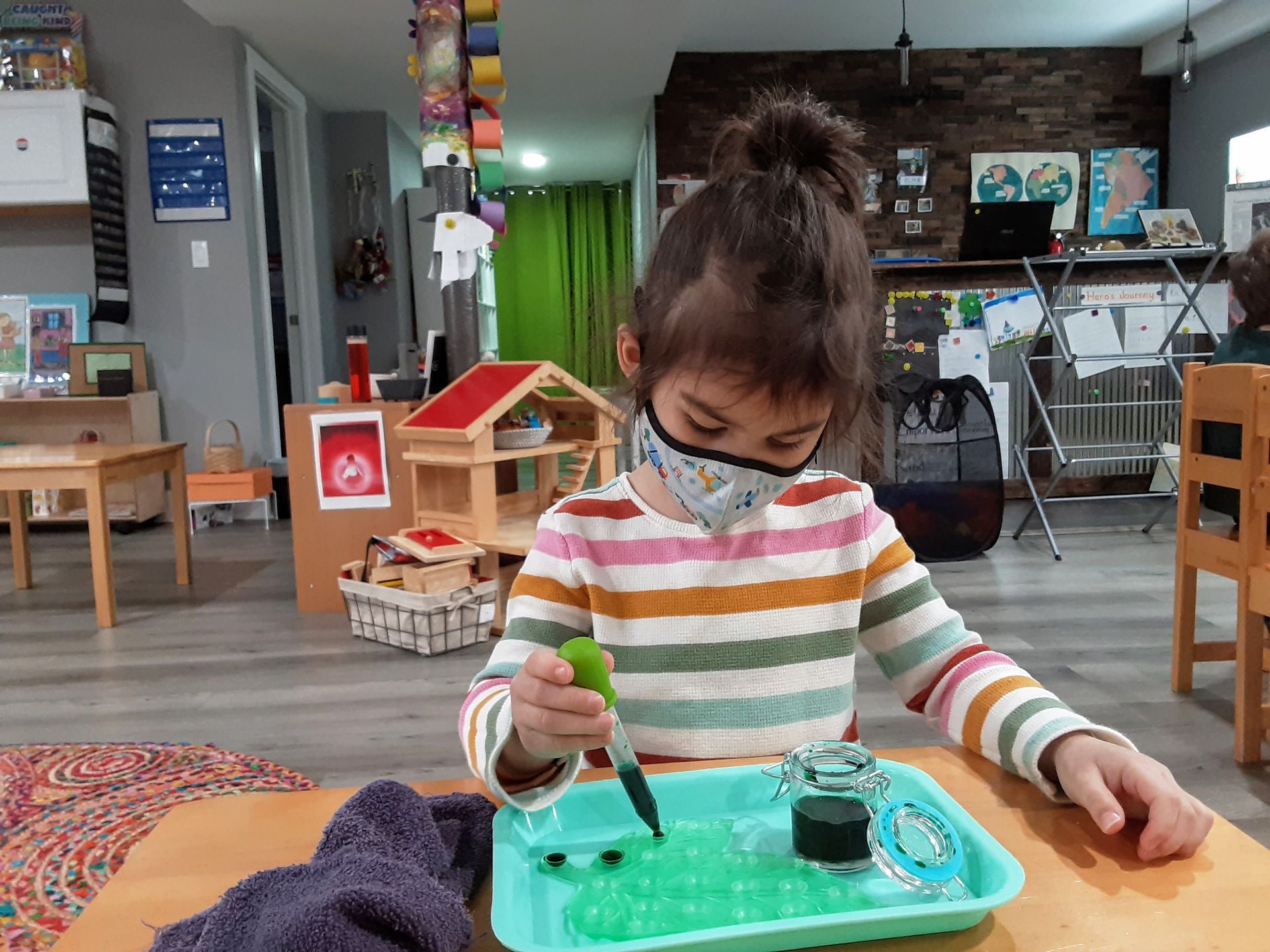 A young child with a facemask is istting at a table dropping droplets of black liquid on a green tray.