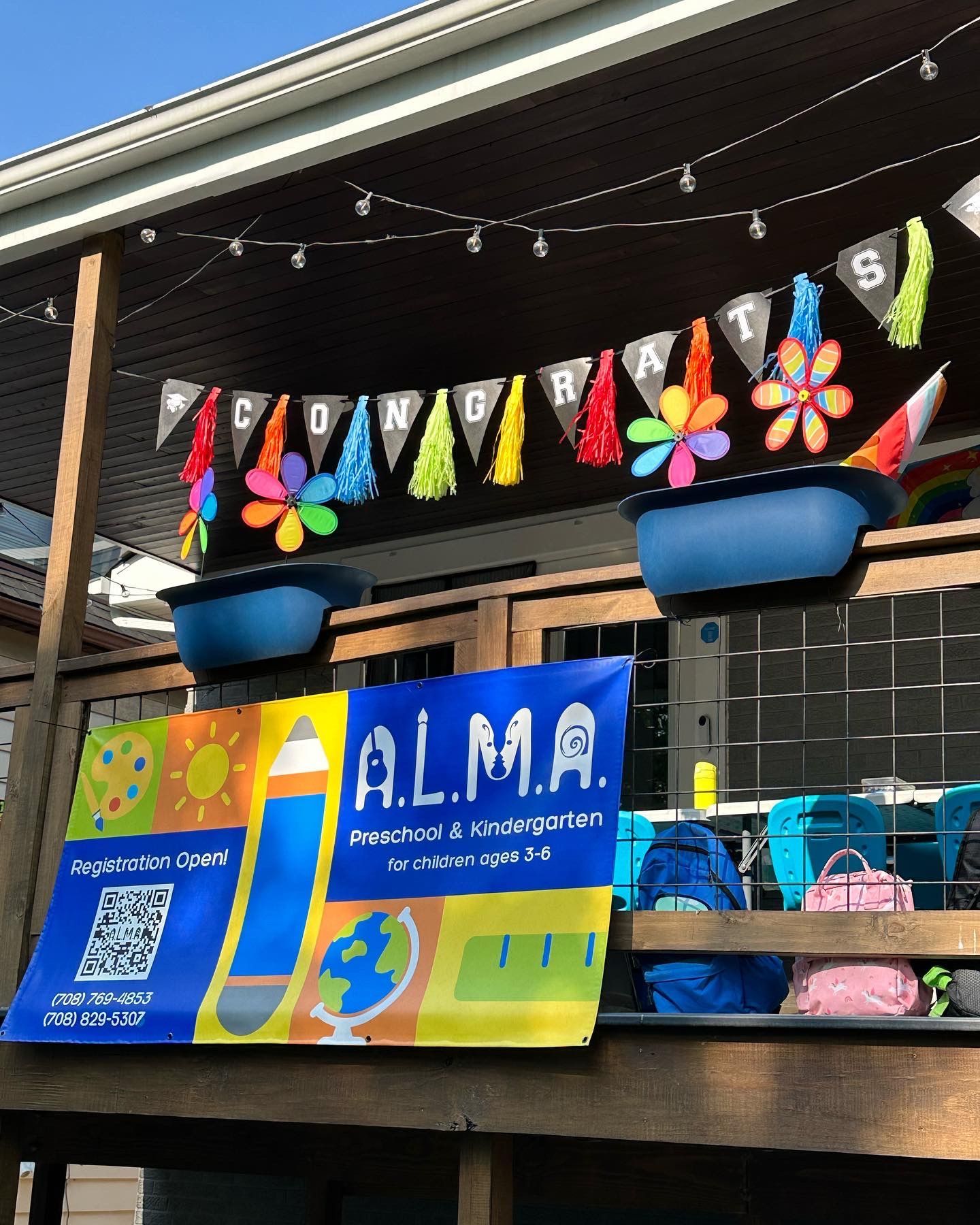 An outdoor porch of a home, deocrated with colorful banners and flags in congratulations. There is a logo of the A.L.M.A. school.