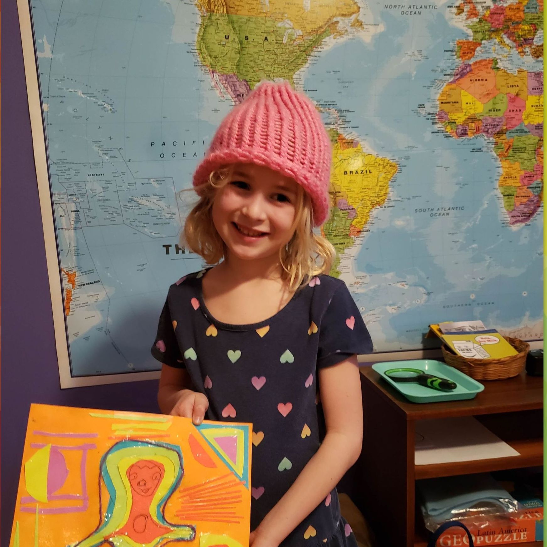 A child with a pink har stand in front of a world map holding a painted picture.