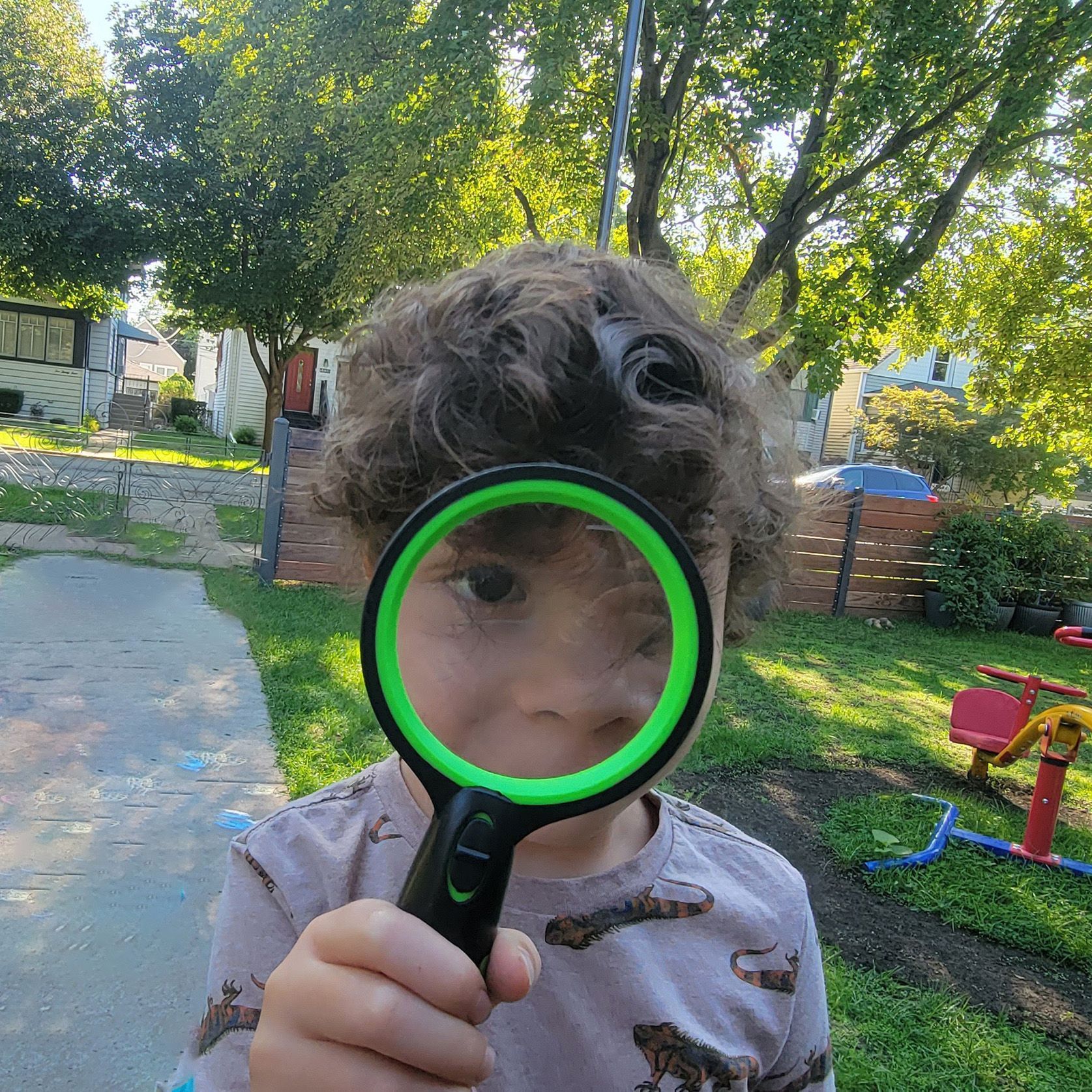A young child peers forward through the lens of a bright green magnifying glass. His face looks larger when seen through the lens.
