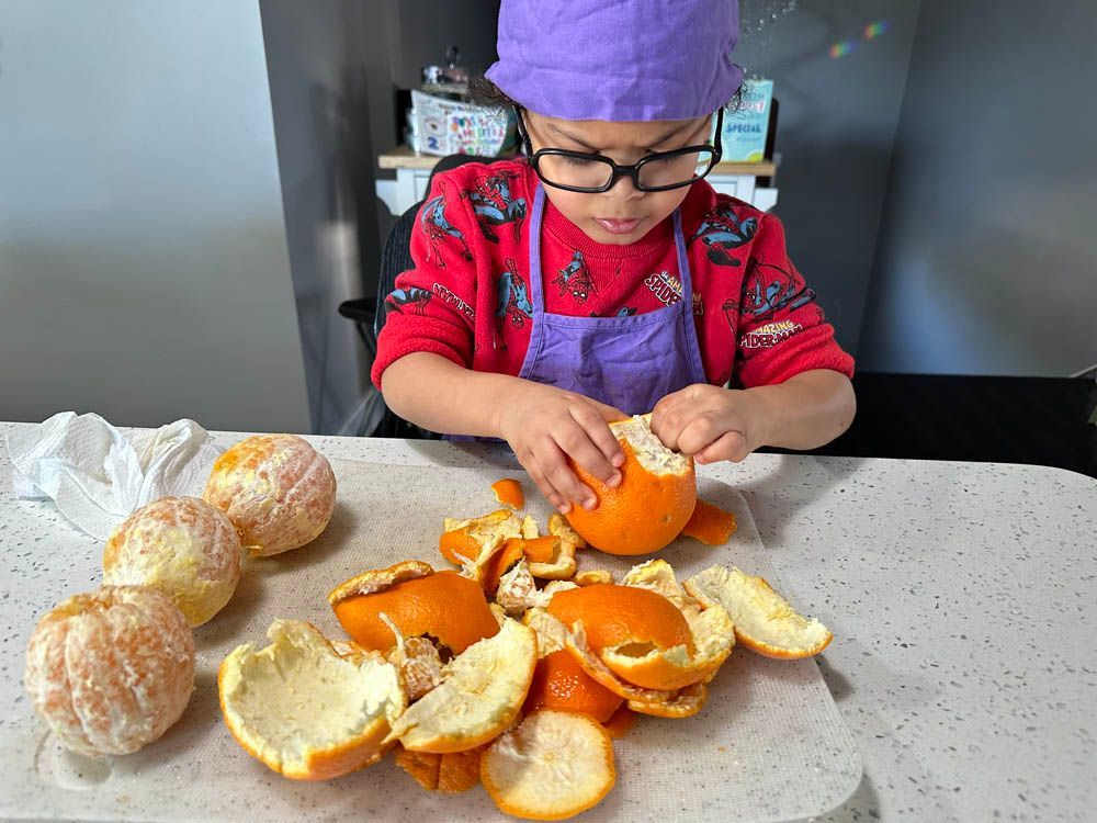 child in purple chef hat and apron peeling oranges at a kitchen table