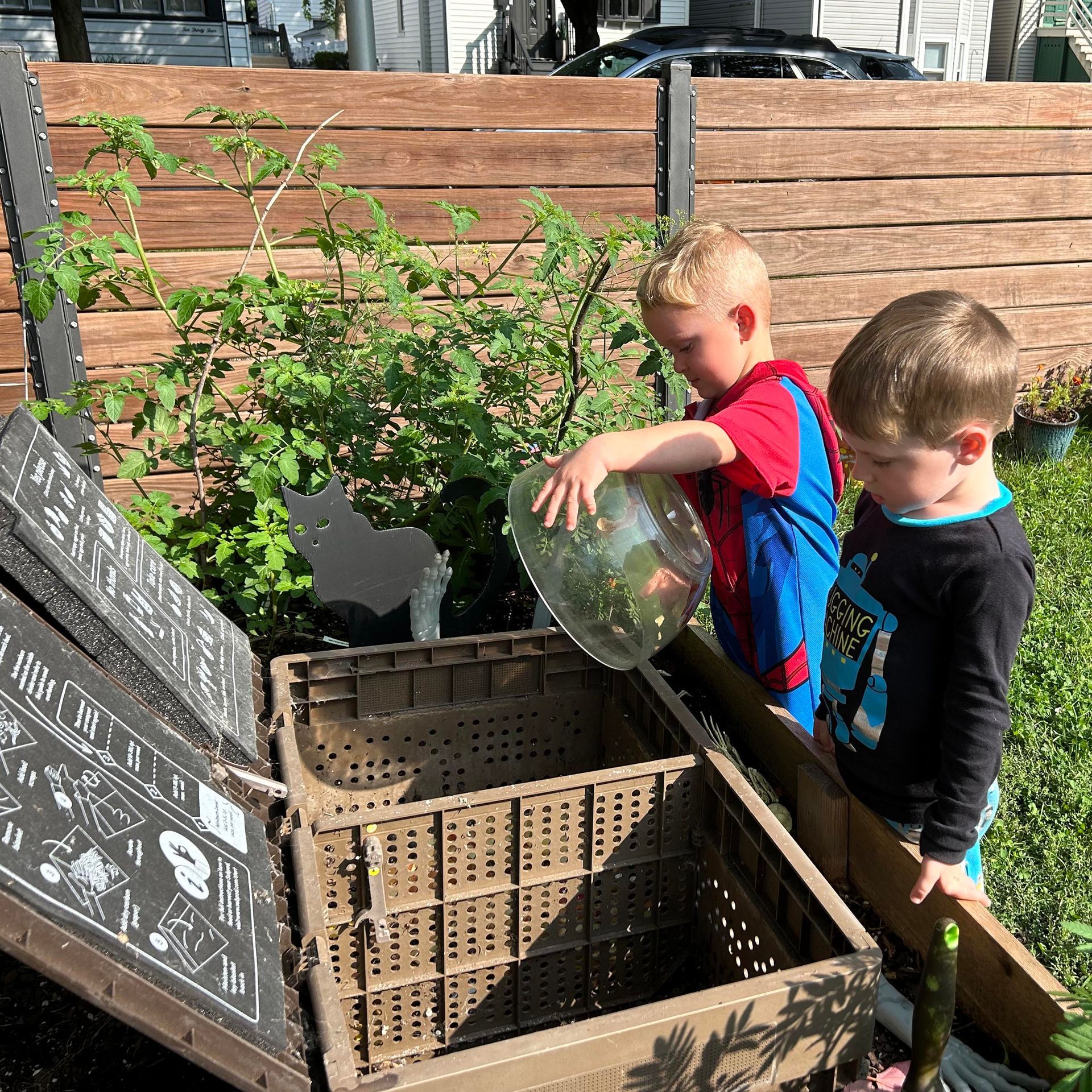 Two children are standing in front of an outdoor compost bin. One child is dumping a large bowl of scraps into the compost bin.