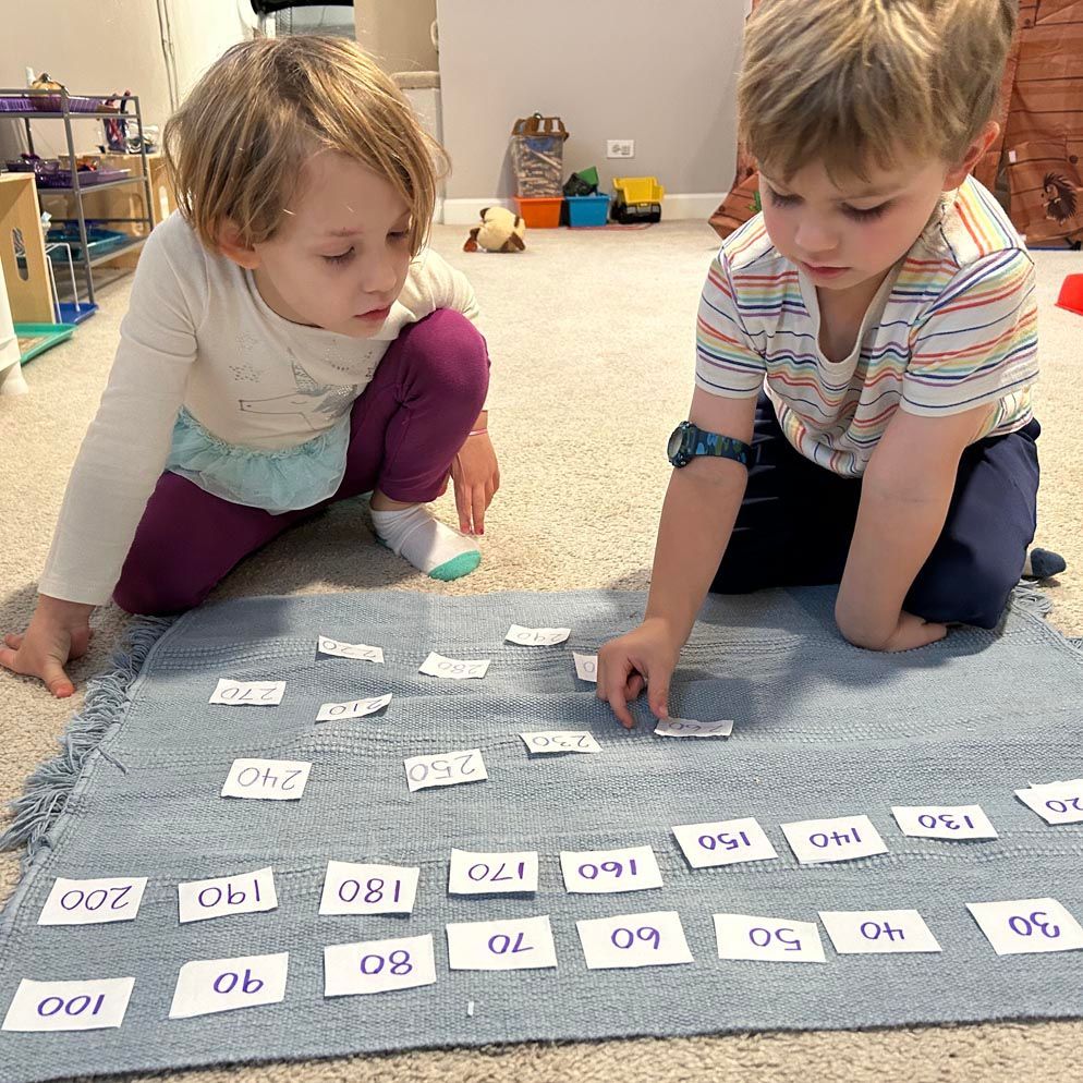 Two young children play with several small white cards that each have a different number. They are arranging the cards on a gray mat.
