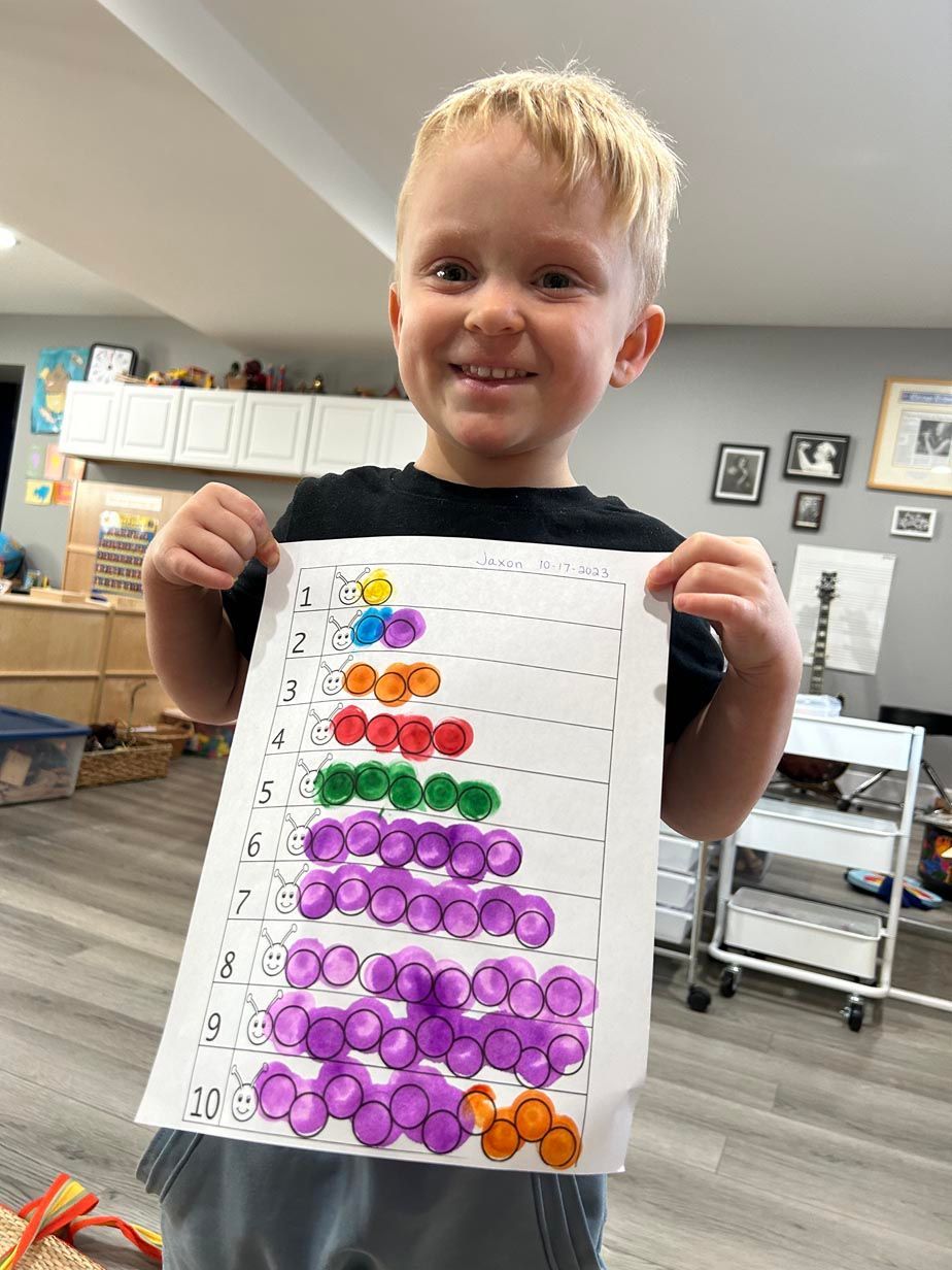 A young child holds up a completed worksheet of primary numbers. Each number is next to a colorful caterpillar that represent each number though a corresponding number of body sections.
