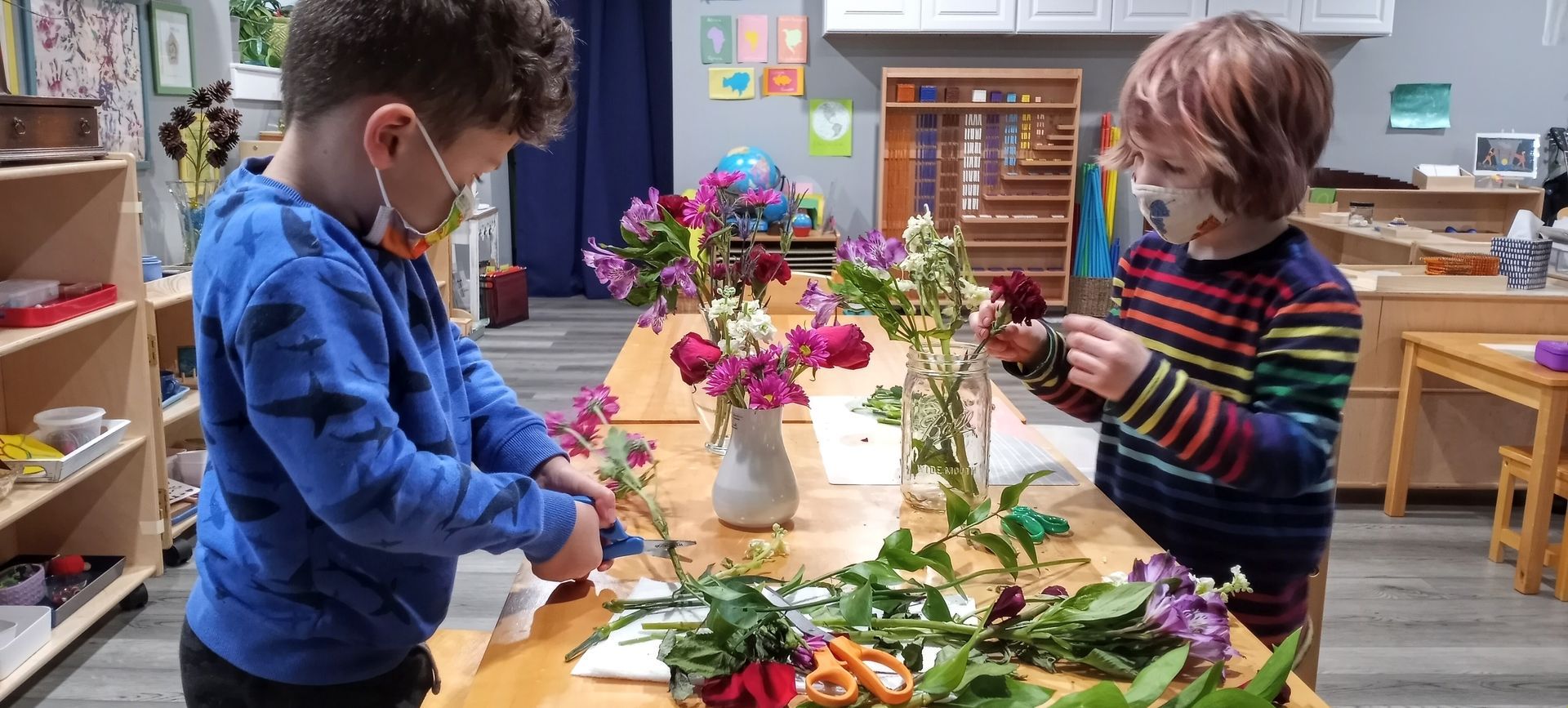 Two children are trimming the stems off flower at a table in a preschool setting, arranging the flowrs in two glass vases.
