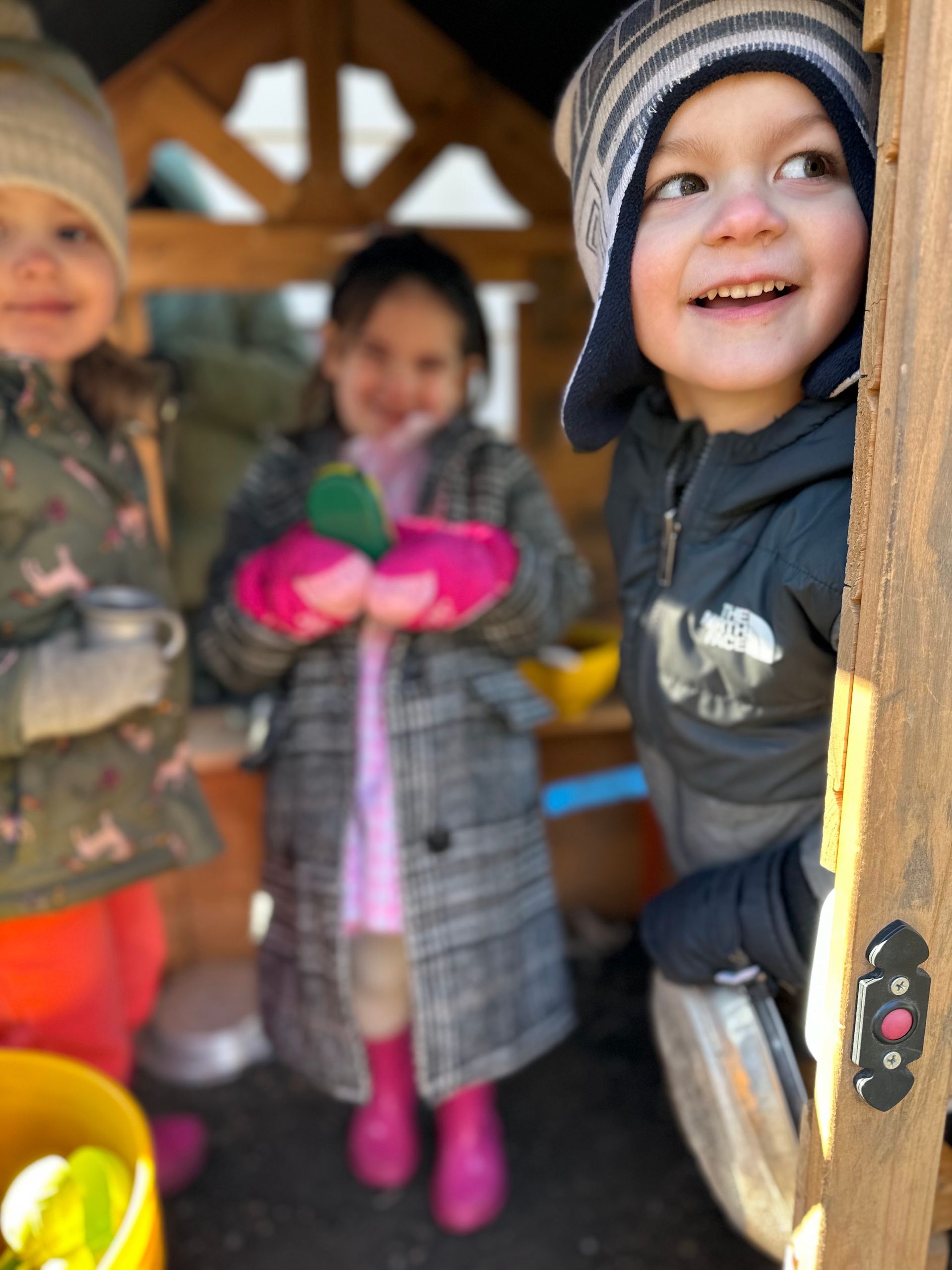 Three young children, dressed in cold weather gear, are gathered indoors at the open doorway of a house, eager to go outside.
