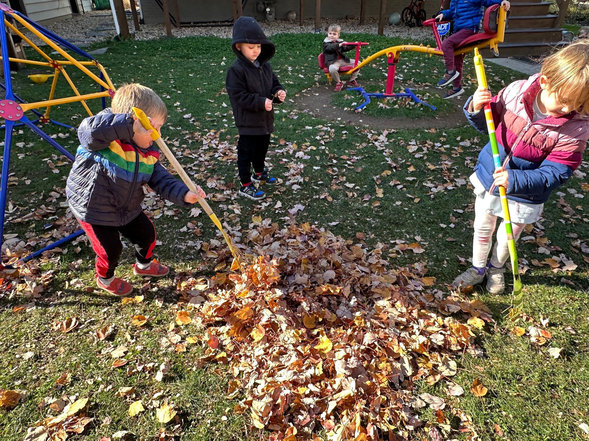 Two young children are raking leaves in a backyard playsetting. There is play equipment in the background and other children on the play equipment.