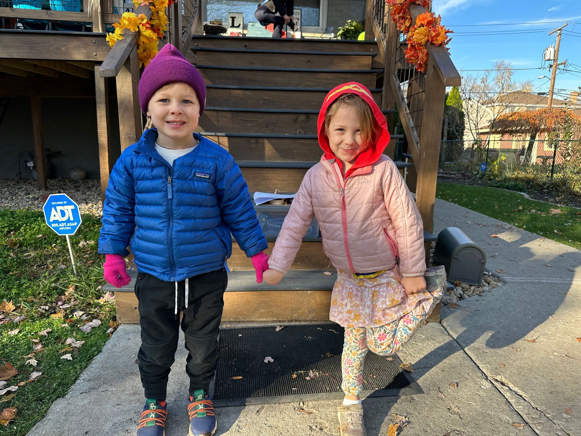 Two small children in winter coats and hats outdoors and holding hands, smiling.