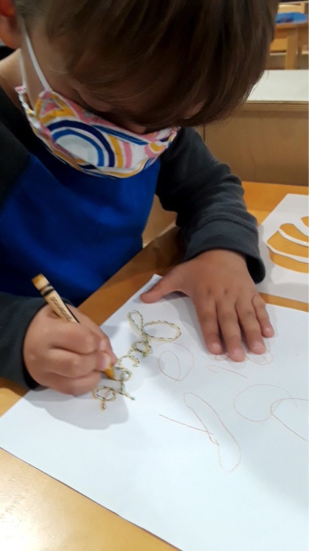 Two hands of a child - one holding a piece of paper down on a table while writing their name with a crayon in the other hand.