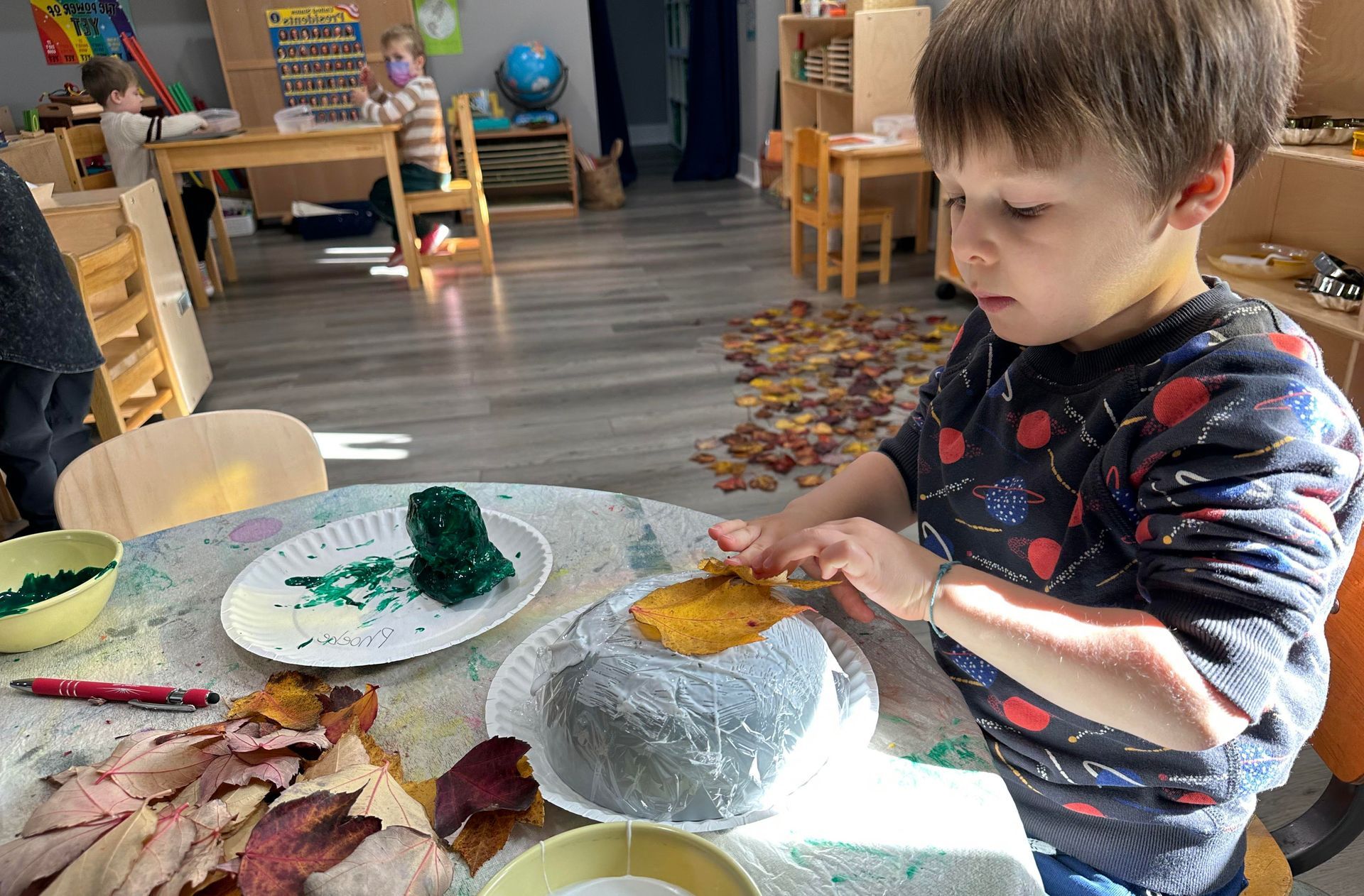 A young child seated at a low table in a preschool setting is working on an art project involving autumn leave pasted to an overturned bowl.