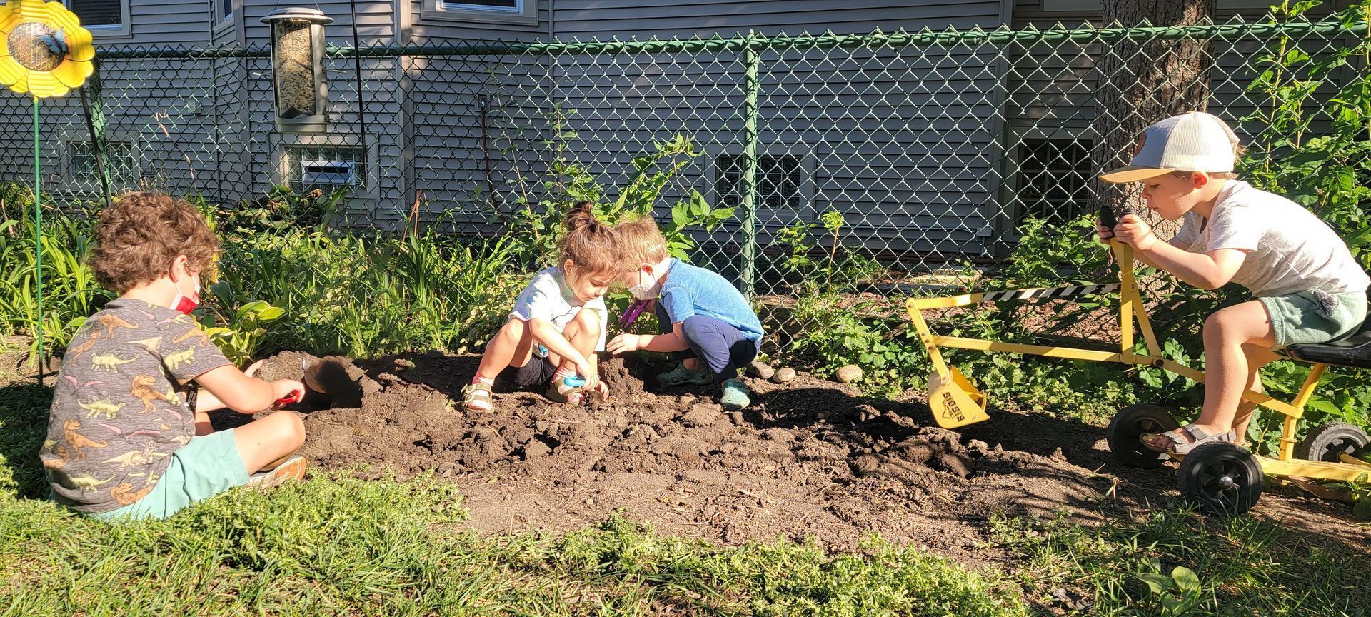 Two young children are digging in a patch of dirt near a chain link fence in a backyard etting.