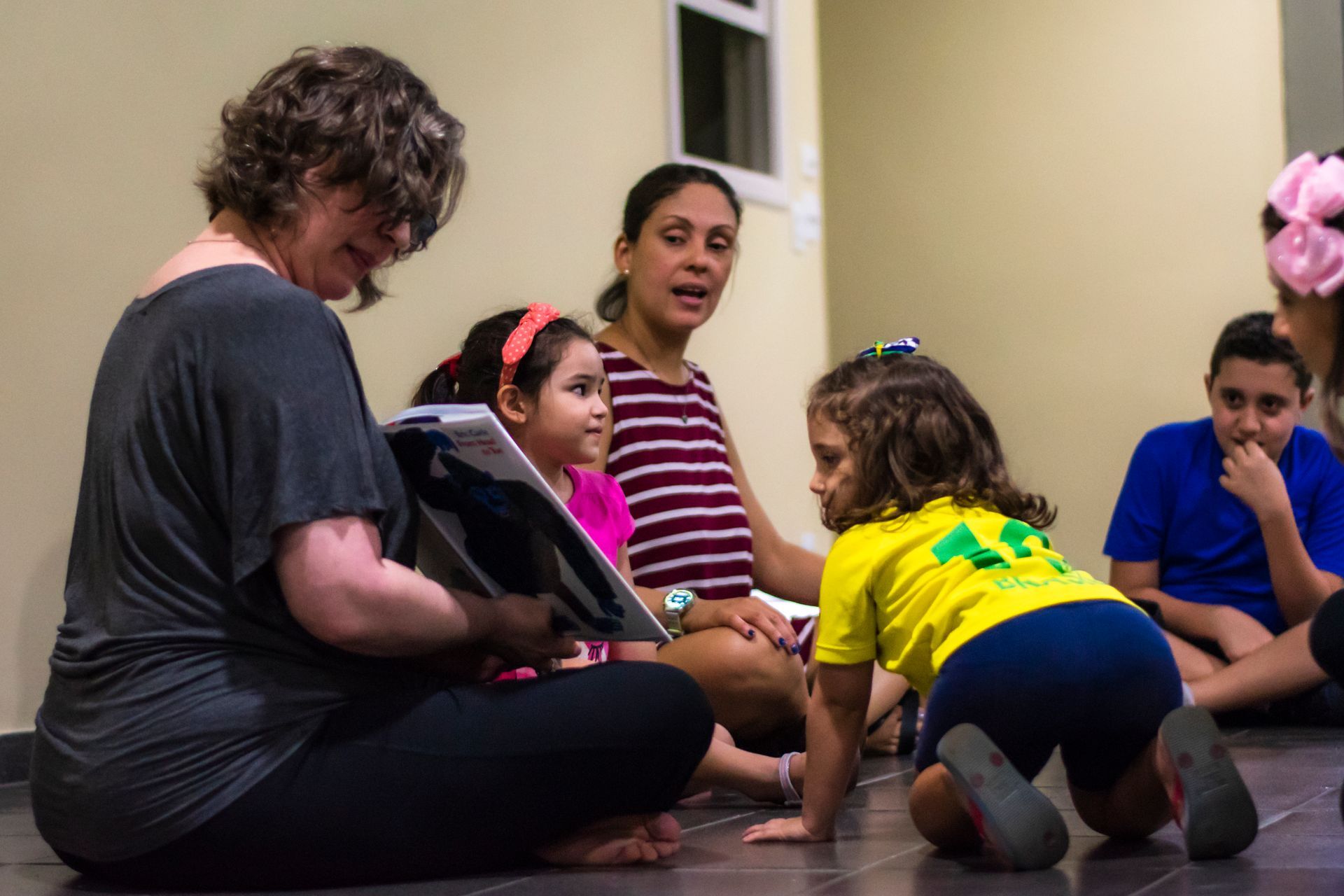 Two teachers seated with a group of children, while one of the teacher reads a storybook. She is showing a picture fron the book to the children.